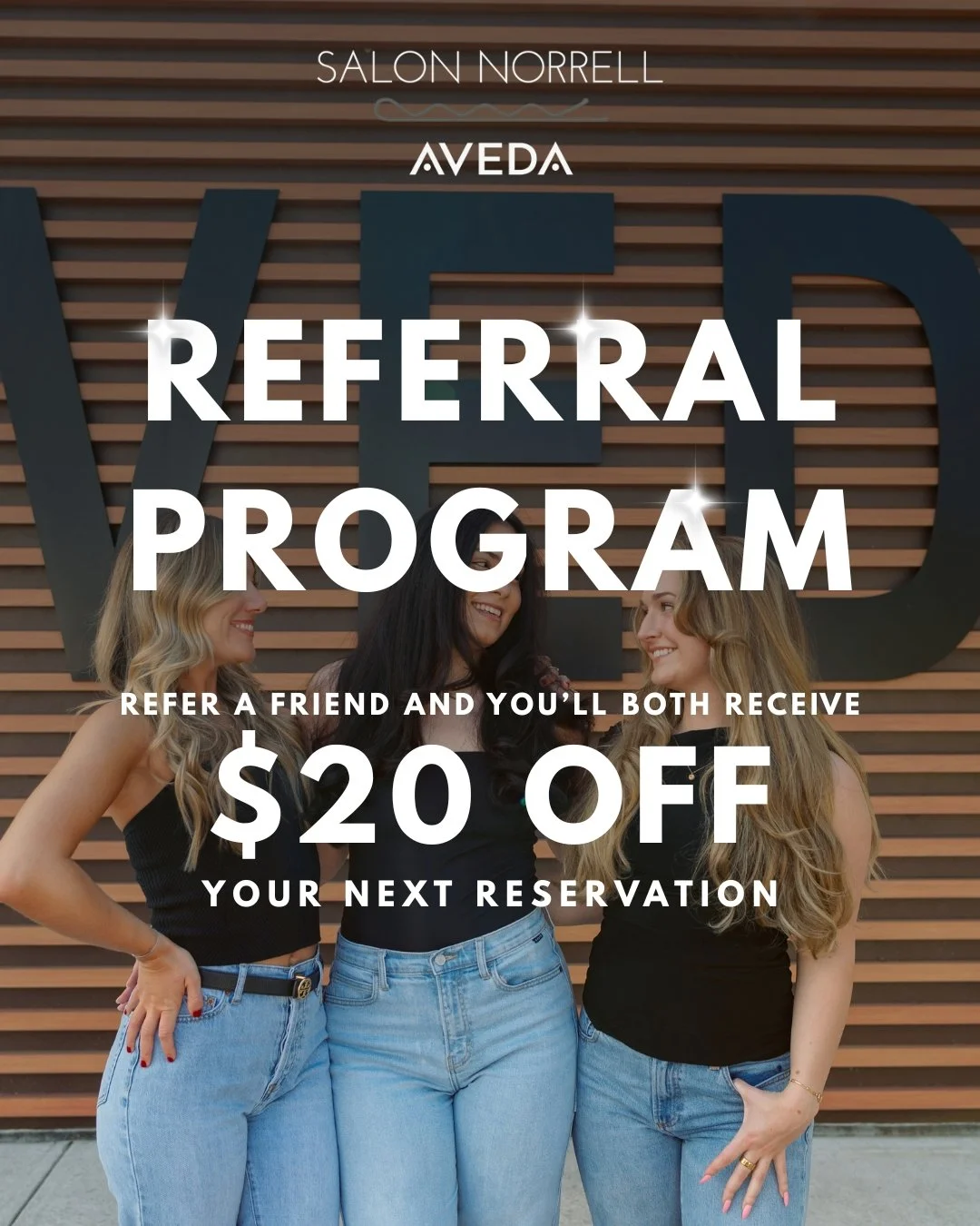 Three women with long hair smiling and talking together, standing in front of a wooden slat wall with the letters 'WED' in large font behind them. The text promotes a referral program offering a $20 discount on the next reservation at Salon Norrell.