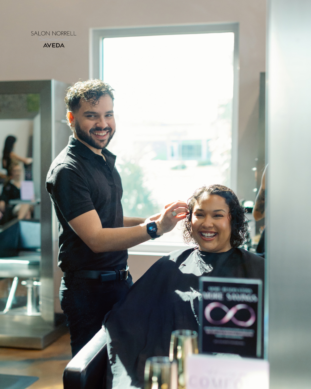 A hairstylist with a beard and black shirt cutting a woman's curly hair at a salon, both smiling. The salon has large windows and contemporary decor.