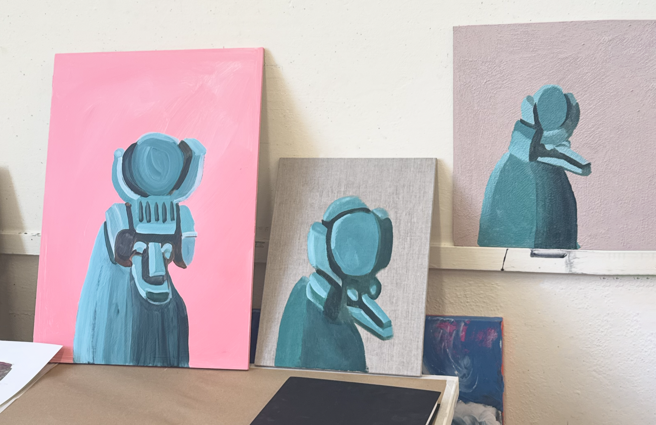 Three paintings of Egyptian deities in shades of blue, pink, and purple, leaning against a wall in an art display.