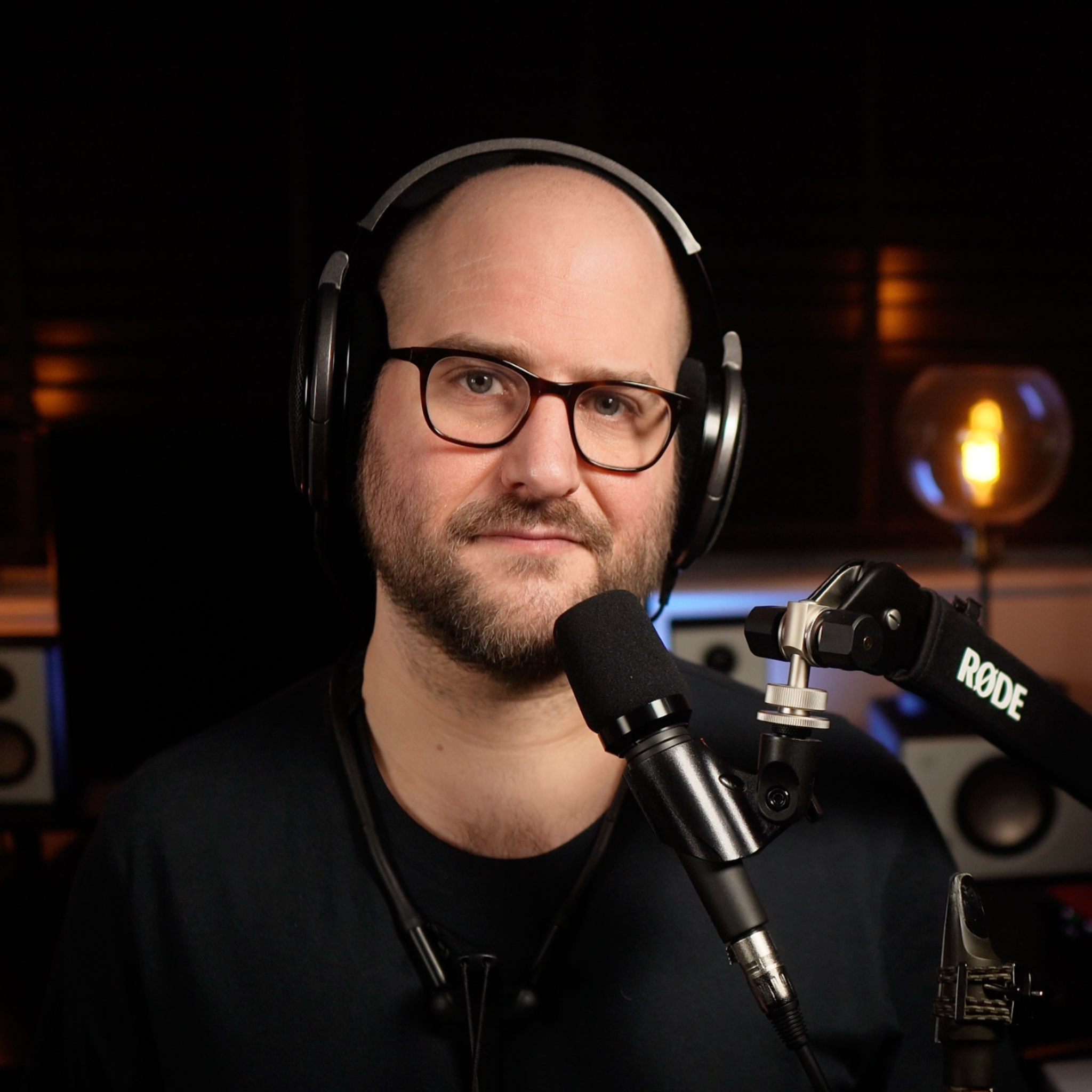 A man with glasses and a bald head wearing over-ear headphones, speaking into a microphone, in a recording studio with dark background.
