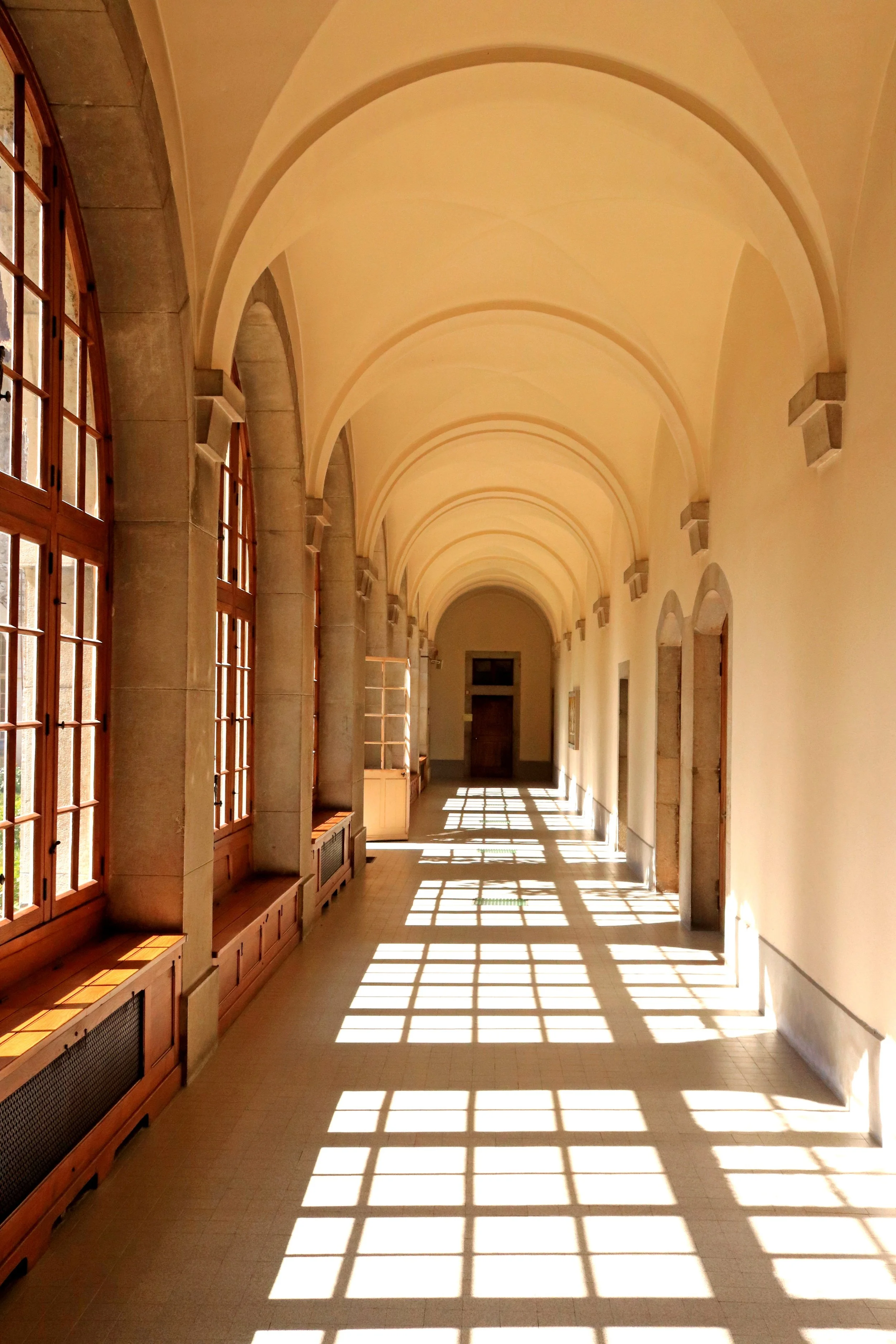 A long corridor with sunlight streaming through large windows on the left, casting shadows on the tiled floor. The ceiling has arched designs, and the walls are painted a light yellow color.