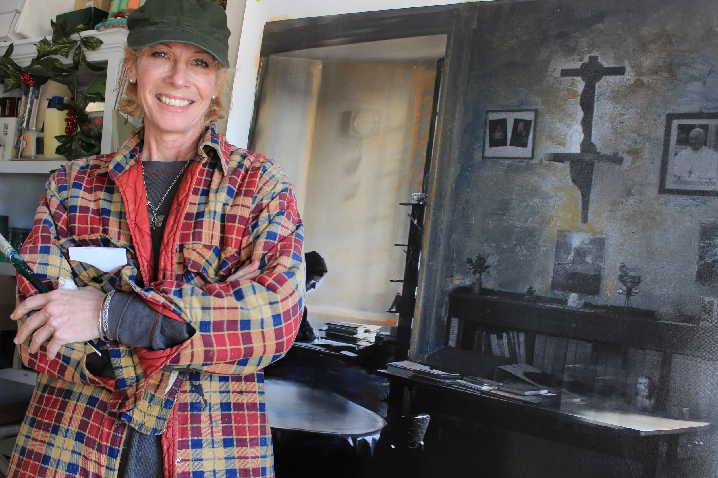 A smiling woman with blonde hair, wearing a green cap and a red and blue plaid shirt, stands in front of a black piano inside a room decorated with Christmas items and framed photos on the wall.
