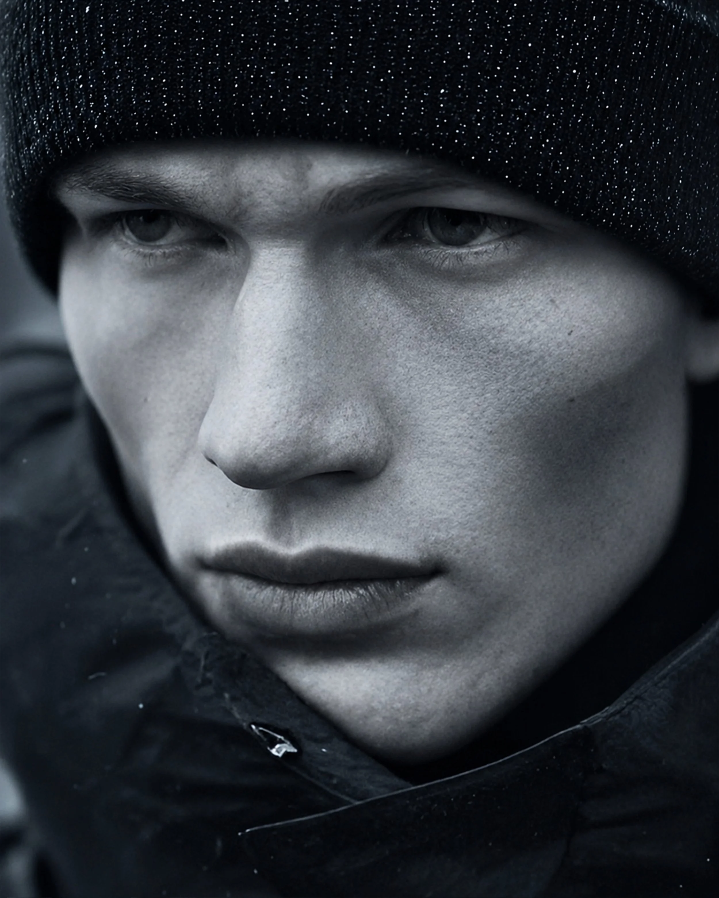 Close-up shot of a person's face partially covered by a black beanie, showing blue eyes, light skin, and a serious expression, with the background and clothing in dark tones.