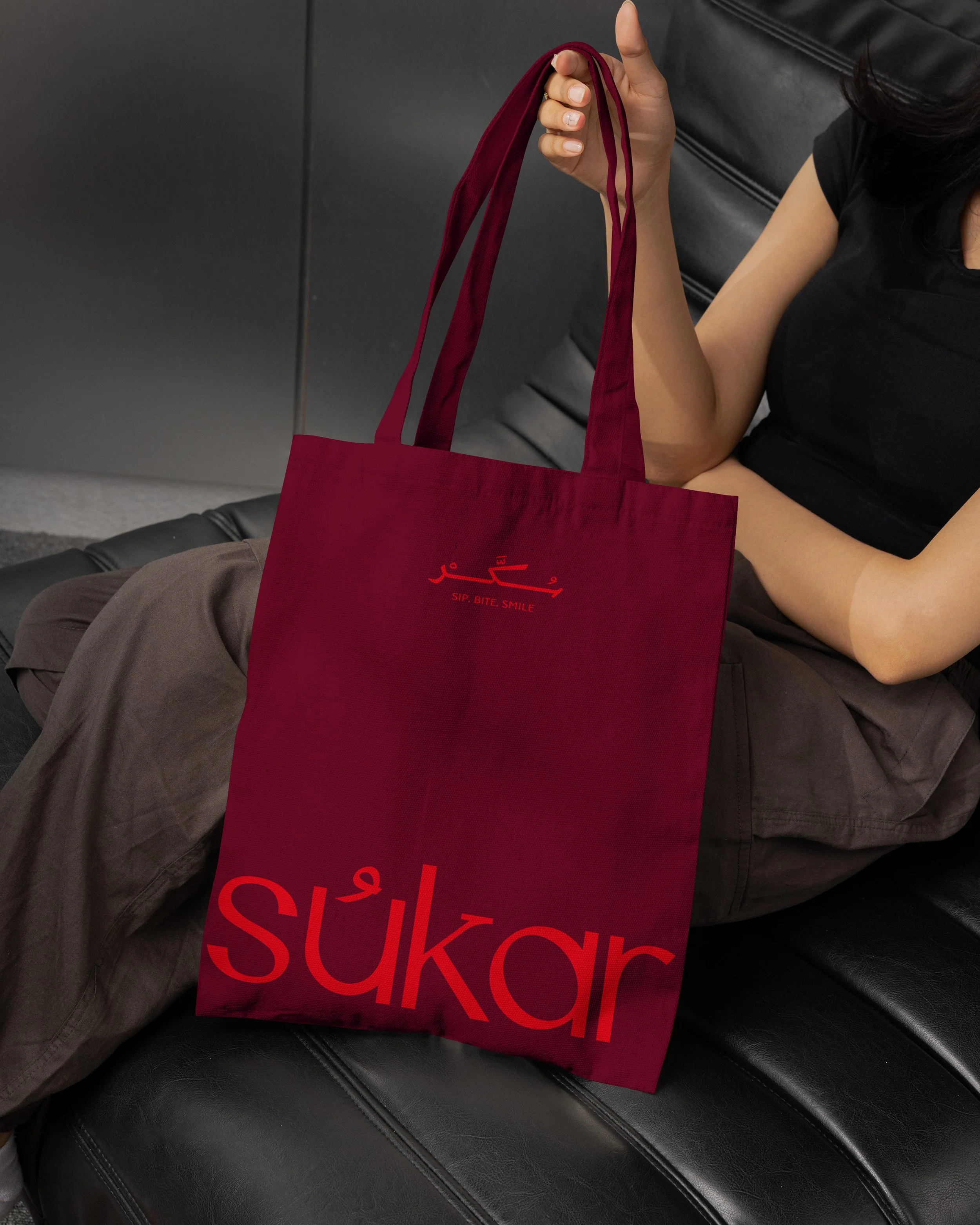 A person seated on a black leather bench holding a red reusable shopping bag with a red logo that reads 'SUKOR' and the words 'SIP. BITE. SMILE' underneath.