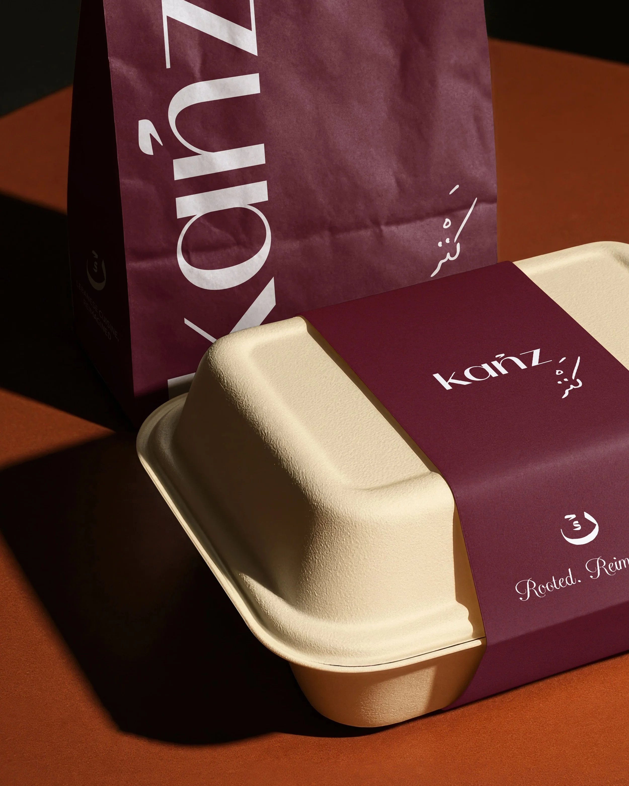 Close-up of a takeout container on a brown surface, with a maroon paper bag featuring white text and logo partially covering the container.