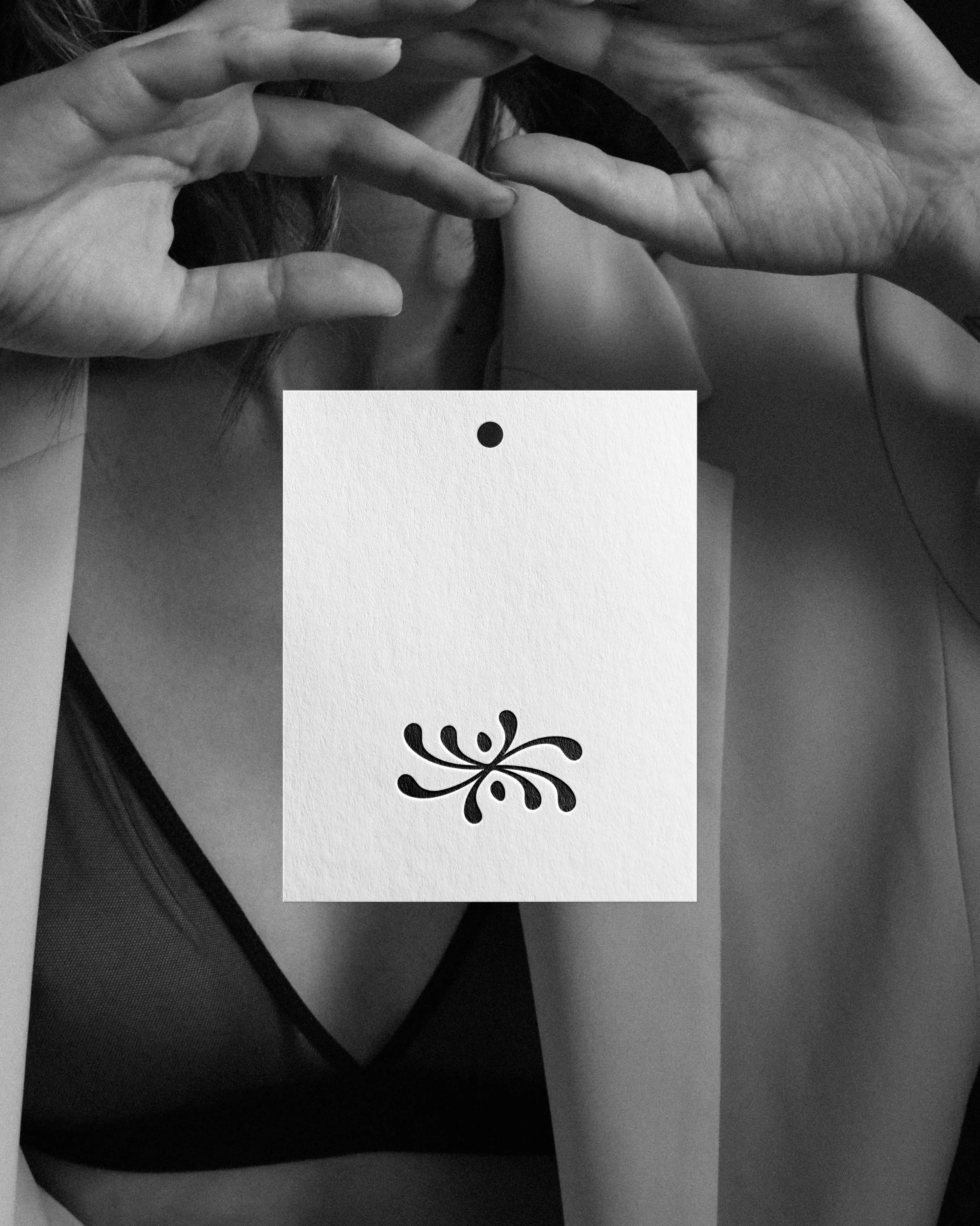 A person holding a phone taking a mirror selfie, wearing a dark jacket and a white card or paper with a black flower or star-like design covering part of their chest. The photo is in black and white.