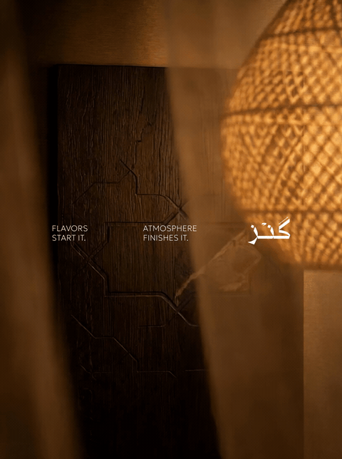 Close-up of a wooden surface with white text and cracks, illuminated by the soft glow of a lamp, with words 'FLAVORS START IT,' 'ATMOSPHERE FINISHES IT,' and a word in Arabic.