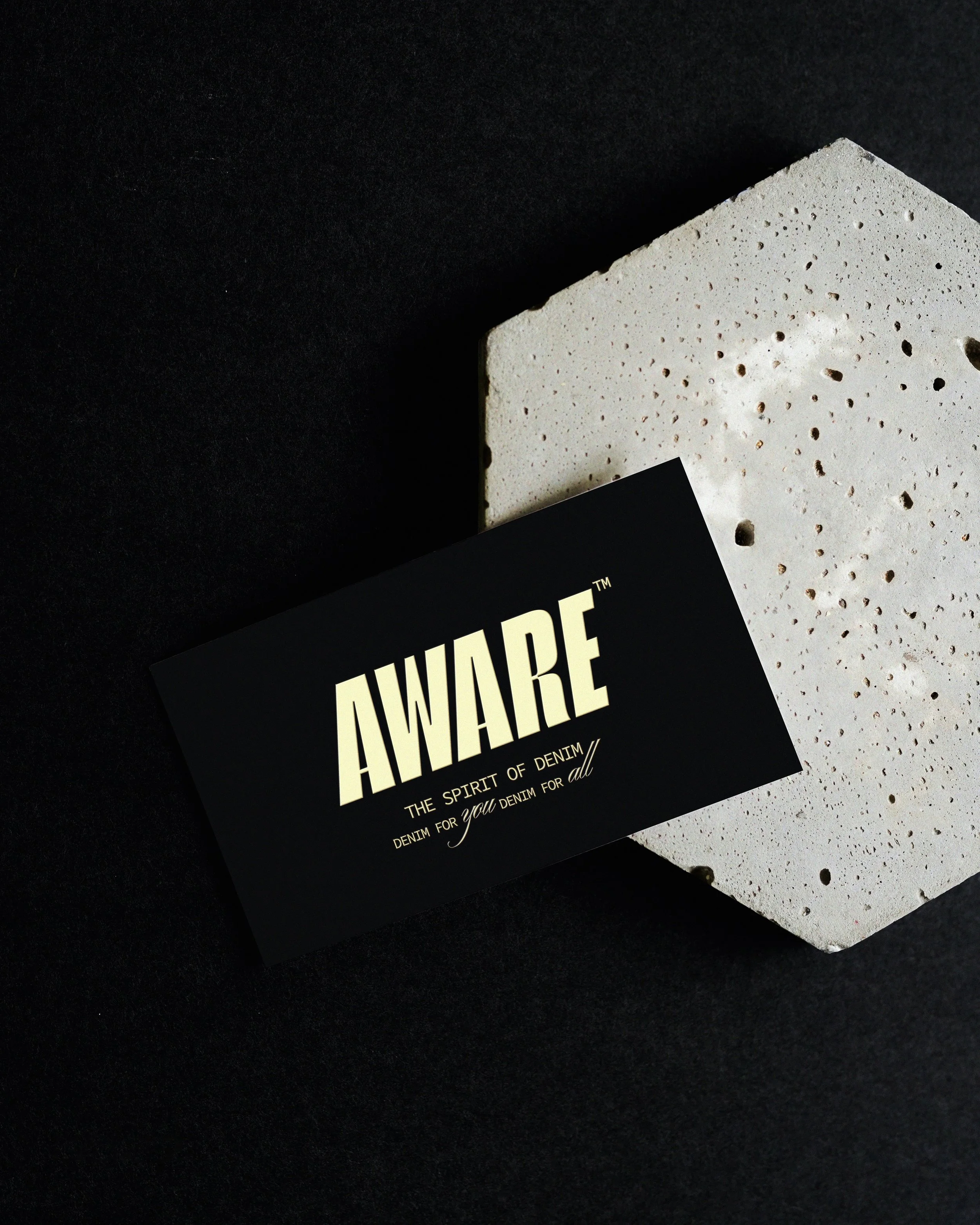 A black card with gold text that reads 'AWARE' and 'The Spirit of Denim for you denim for all,' placed on a speckled white surface with a black background.