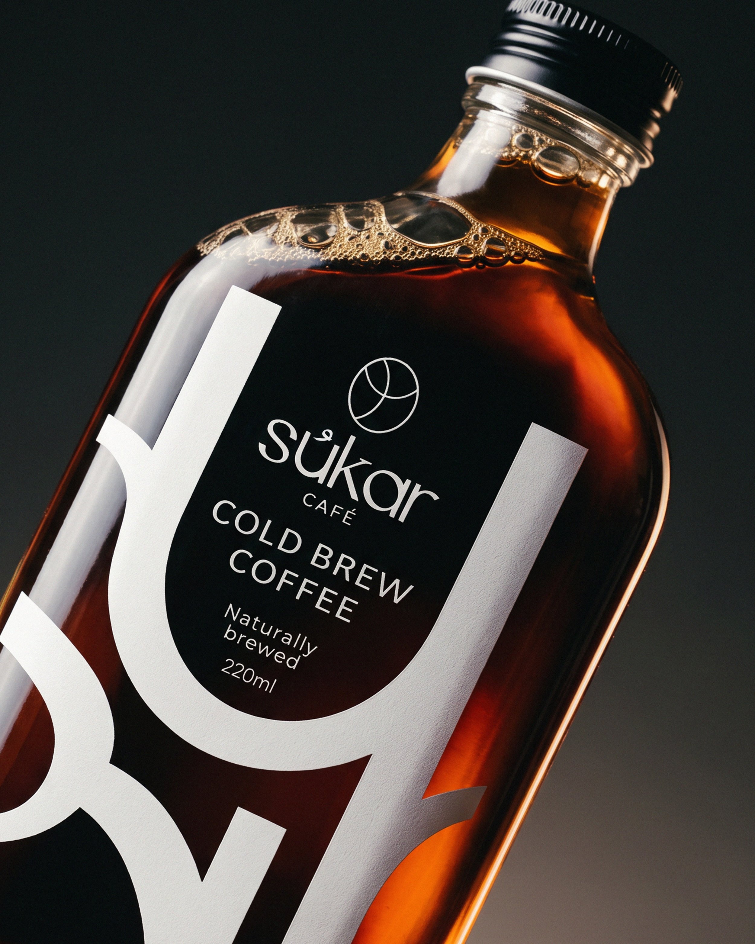 Close-up of a brown glass bottle with a black screw cap, containing cold brew coffee. The label reads "Sukkar Café Cold Brew Coffee, Naturally Brewed, 220ml." The background is dark, highlighting the bottle and bubbles inside the coffee.