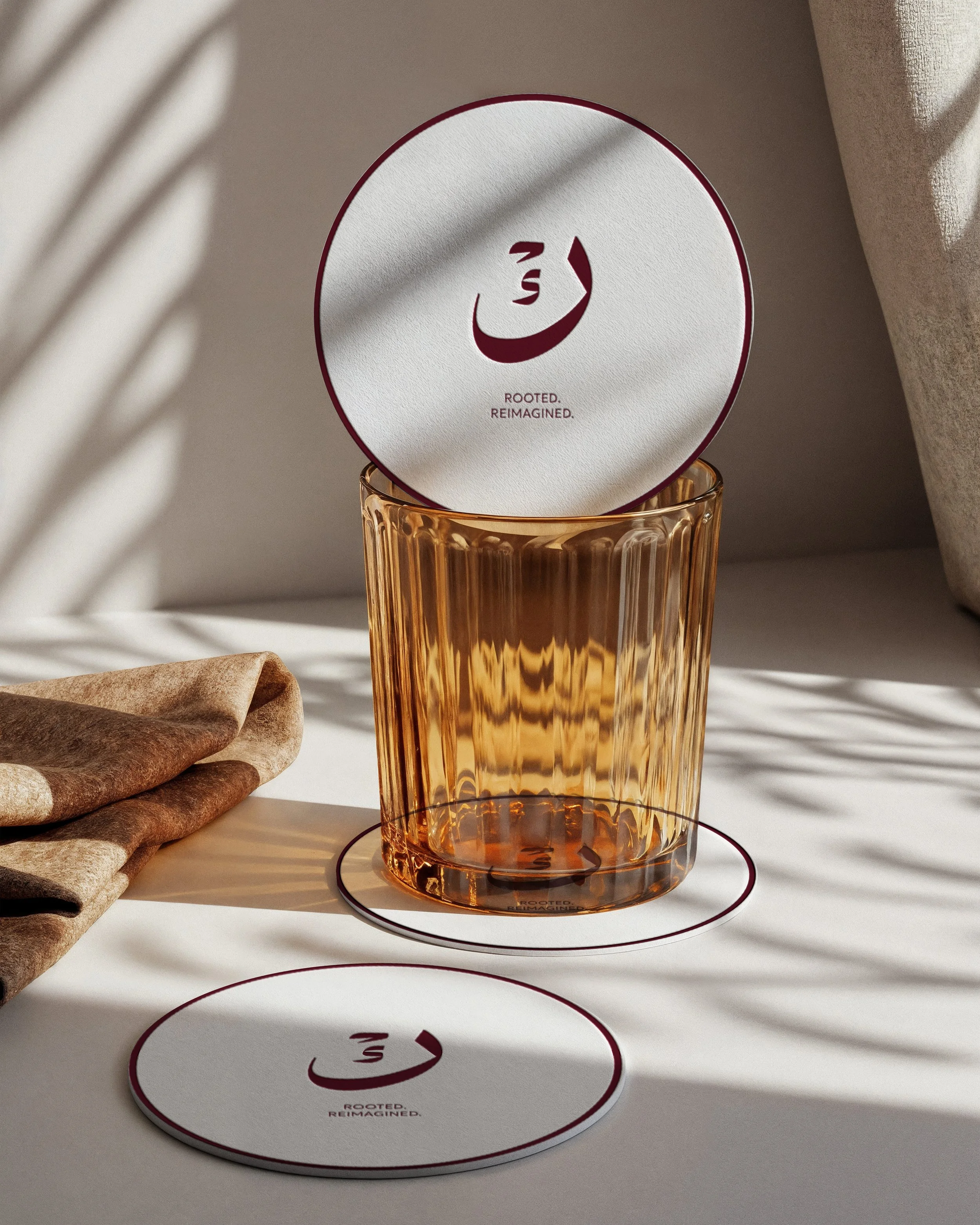 A round mirror with a maroon border and a face logo with closed eyes and a smile, with the words 'Rooted, Reimagined' underneath, displayed above a tall amber-colored glass, on a white surface with shadows and sunlight.