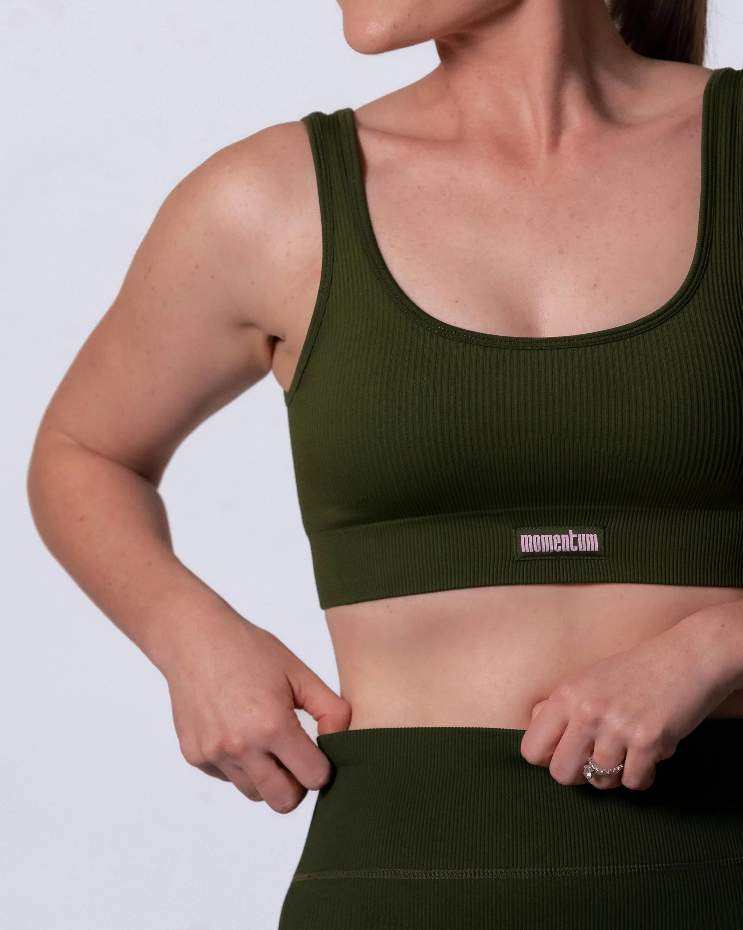 Close-up of a woman wearing an olive green sports bra with a pink and black label reading "momentum". She has reddish hair tied back, and her face is not fully visible. She has freckles and her arms are resting on her hips, with her left hand near he