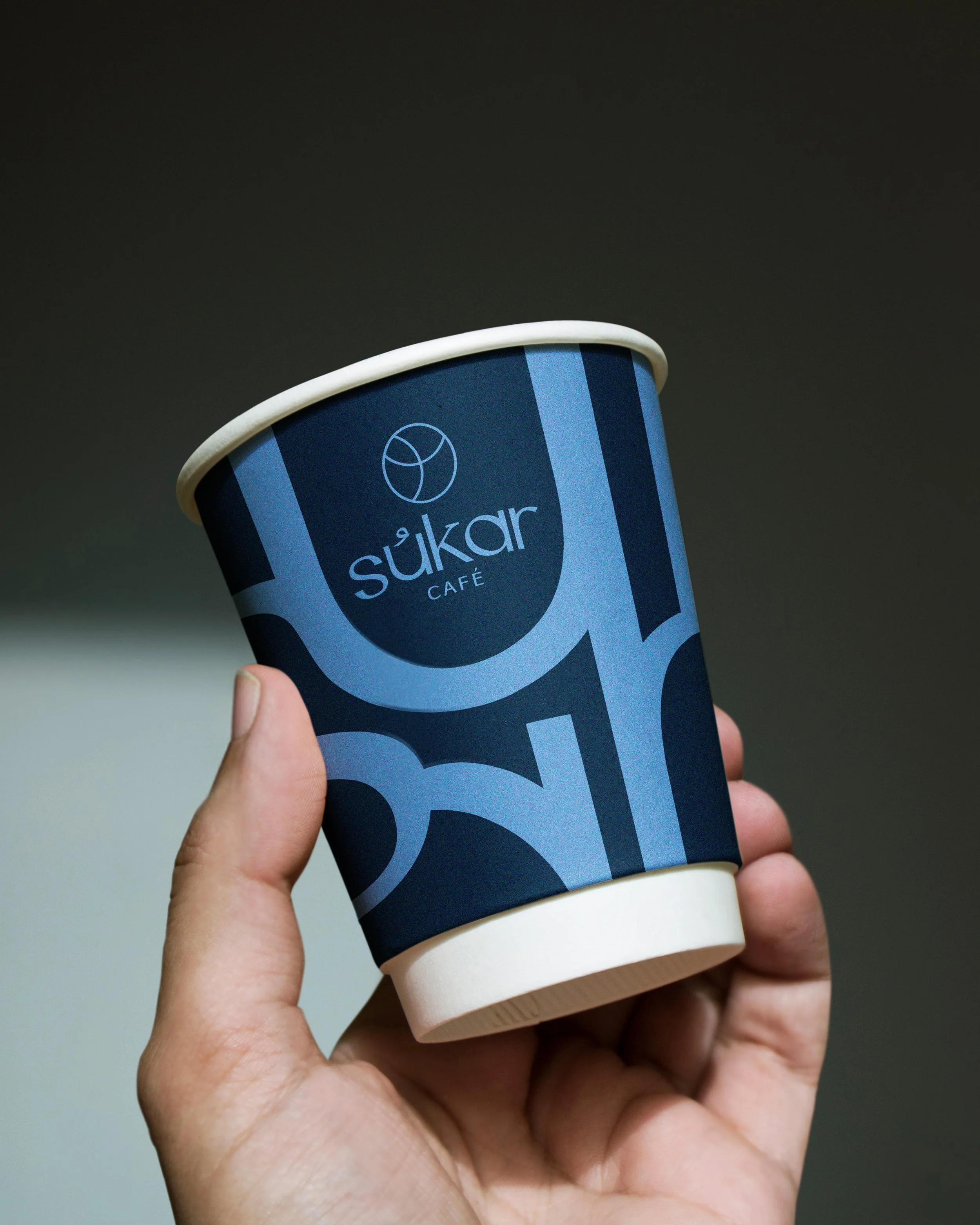 A hand holding a paper cup with a blue sleeve that says 'sükar CAFE' and a white plastic lid.