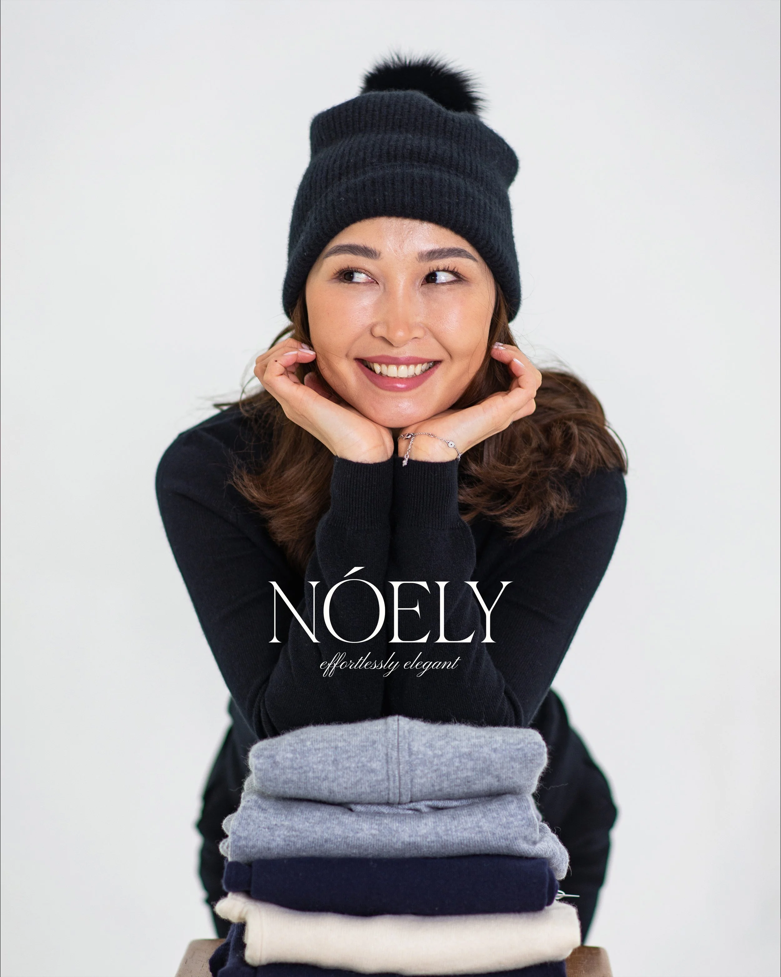 A woman with long curly hair wearing a black beanie with a pom-pom and a black sweater, smiling and resting her chin on her hands, stands behind a stack of folded clothing in various colors and fabrics against a plain white background. The text 'NÖELY effortlessly elegant' is overlaid on the image.