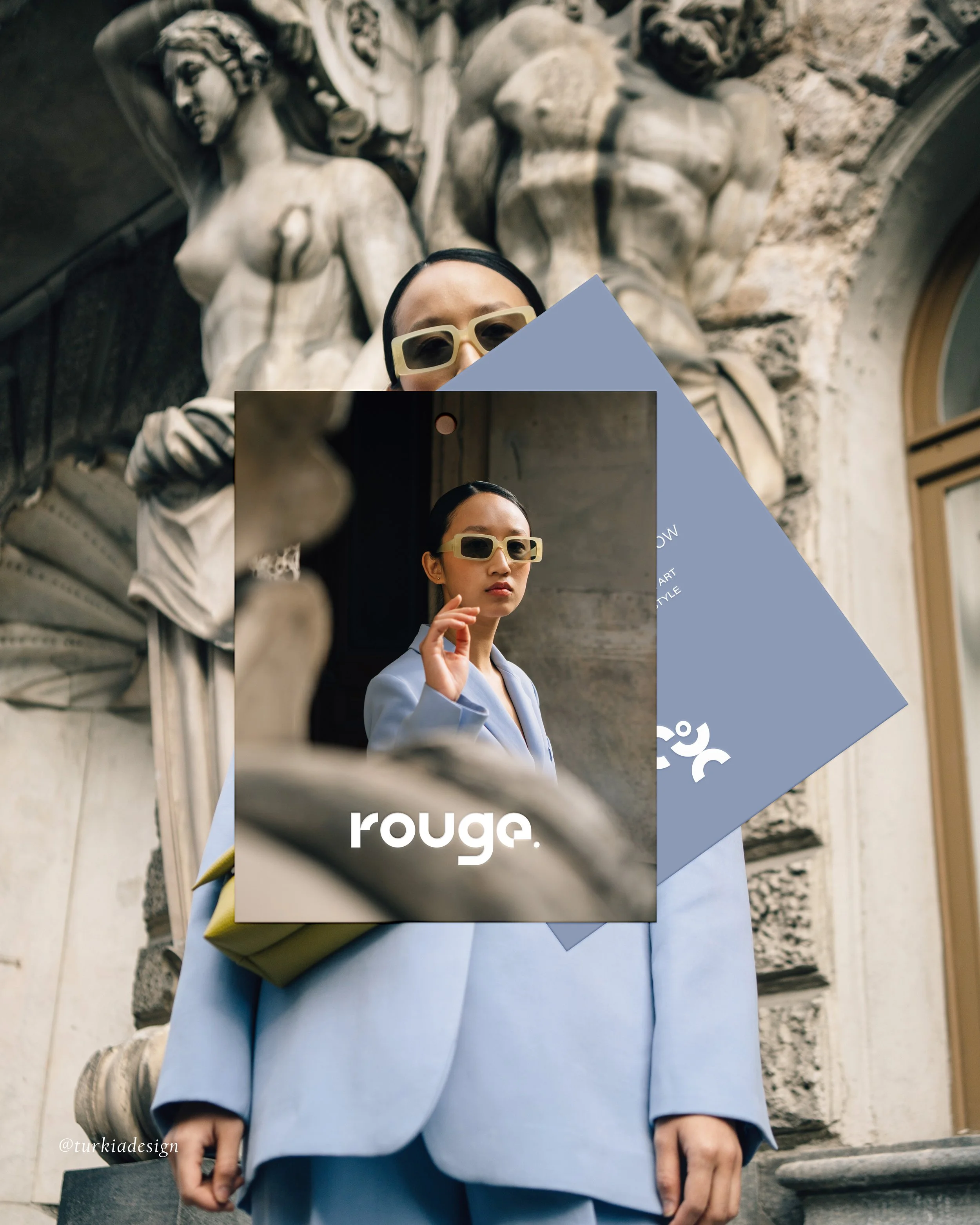 A woman wearing large beige sunglasses, a light blue blazer, and a matching light blue dress standing in front of a sculpture of muscular figures carved into a stone wall, holding a beige purse with a yellow handle.