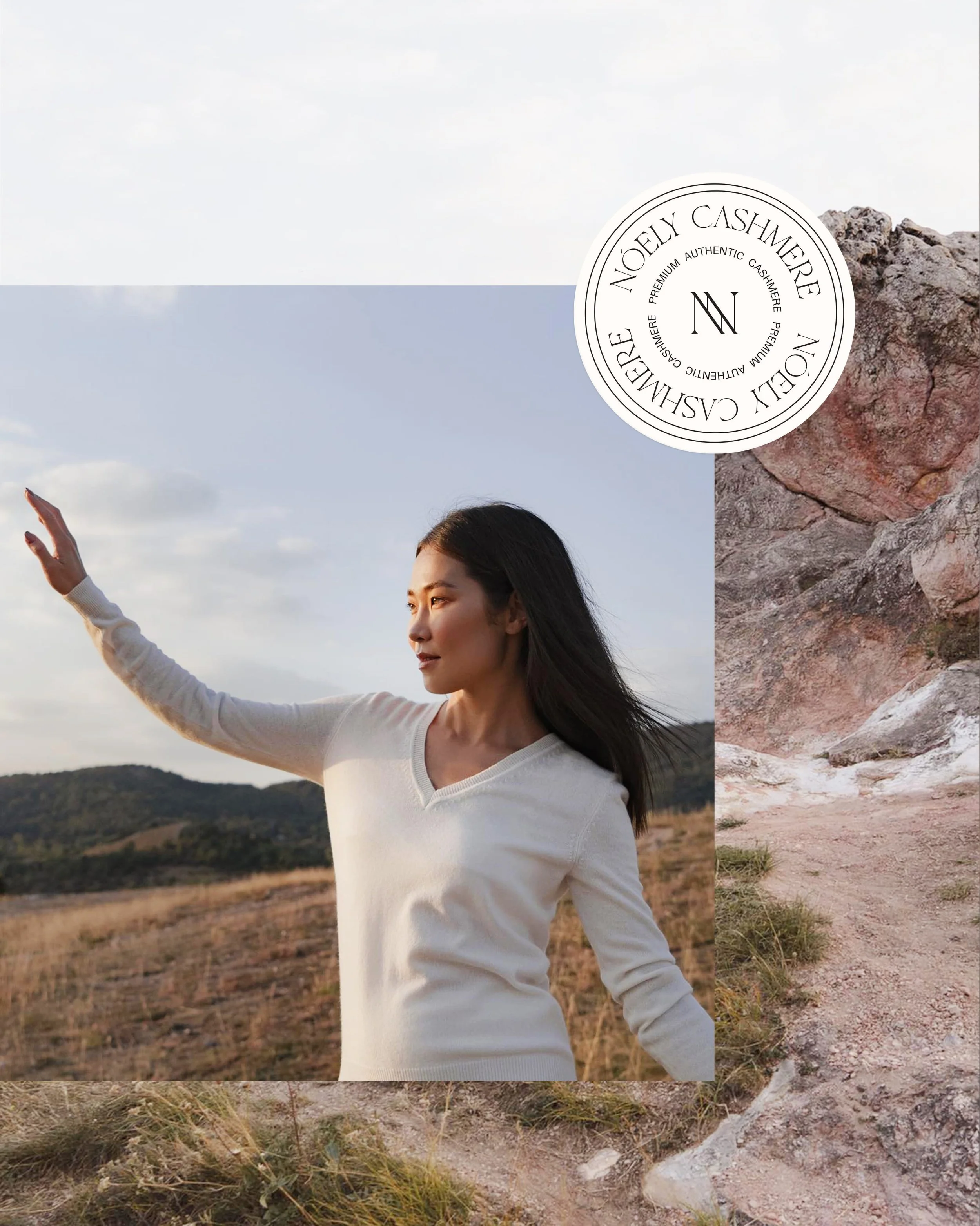 A woman wearing a white long sleeve shirt standing outdoors in a field with mountains in the distance, raising her right arm as if reaching for or pointing at something, with a logo overlay reading 'Noely Cashmere' and additional text about premium a