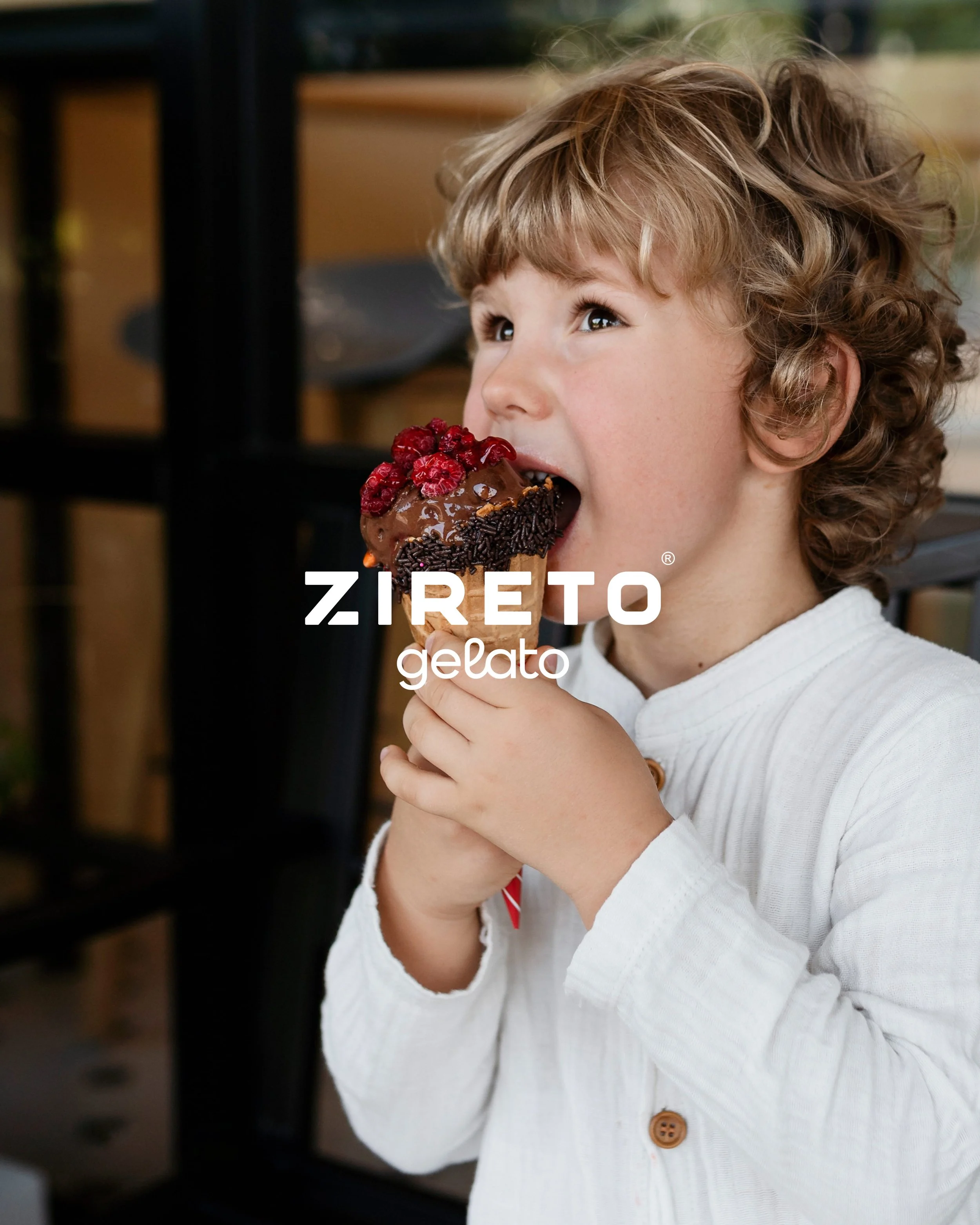A young child with curly hair and light skin holding a chocolate ice cream cone topped with raspberries and chocolate sprinkles, wearing a white long-sleeve shirt with a brown button.