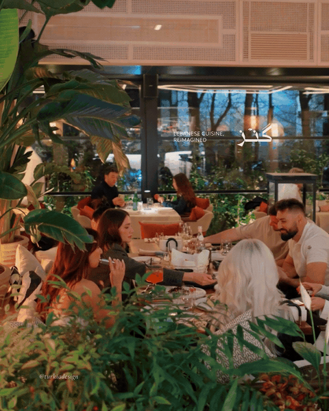 People dining inside a restaurant with large windows and lush green plants.