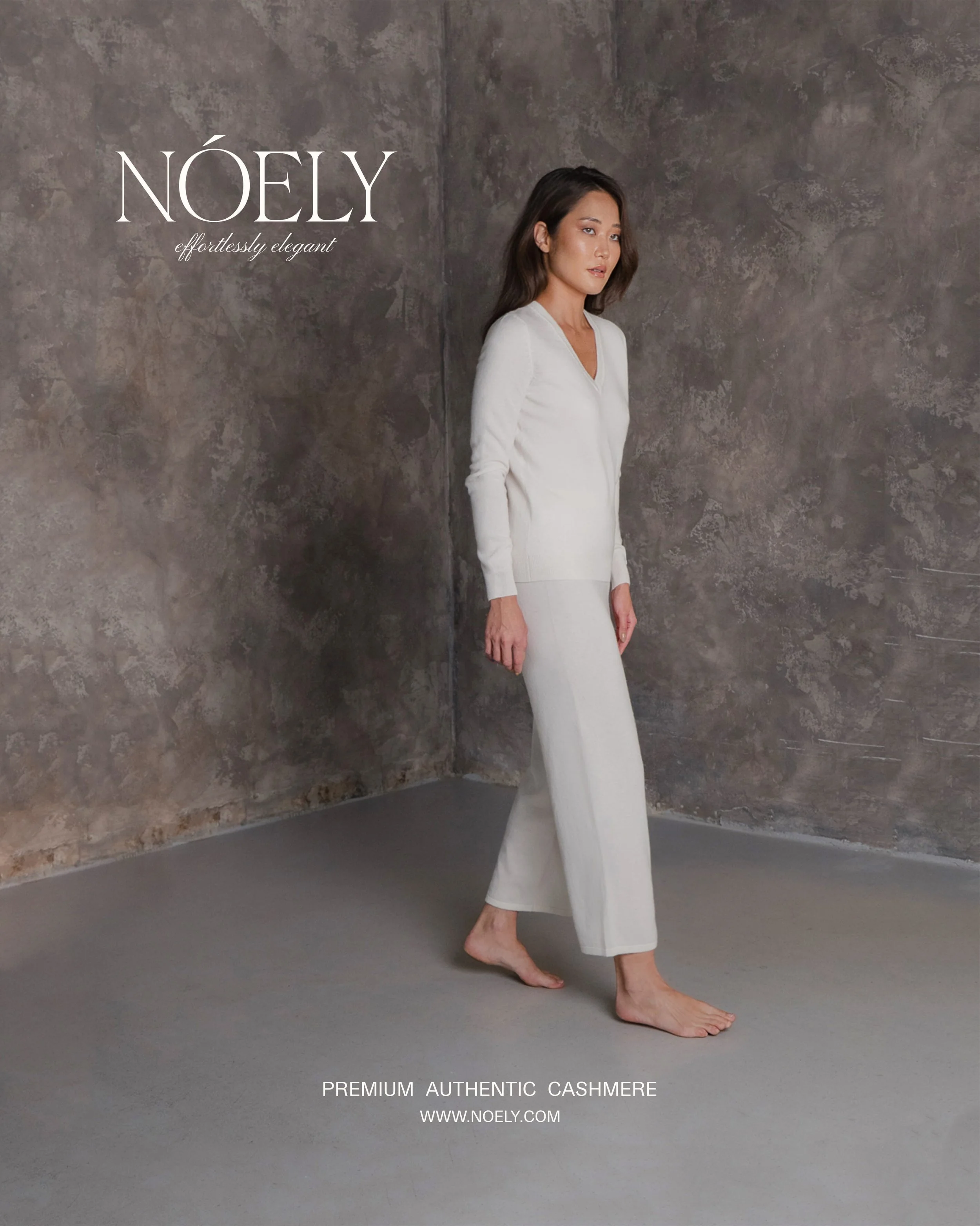 A woman wearing a white cashmere outfit standing against a textured gray background with text promoting NOELY premium authentic cashmere and website at the bottom.