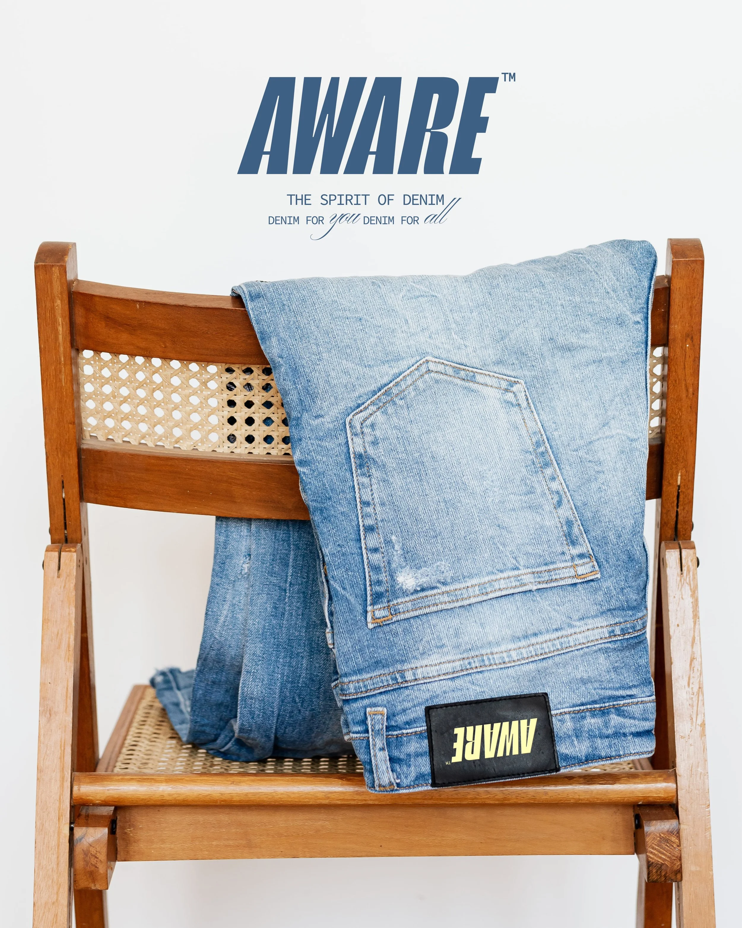A wooden chair with a cane backrest and a denim jacket draped over the backrest. A pair of jeans is folded on the seat, and a black rectangular badge with the brand name AWAYE is partly visible on the lower part of the seat. Above the chair, on the w