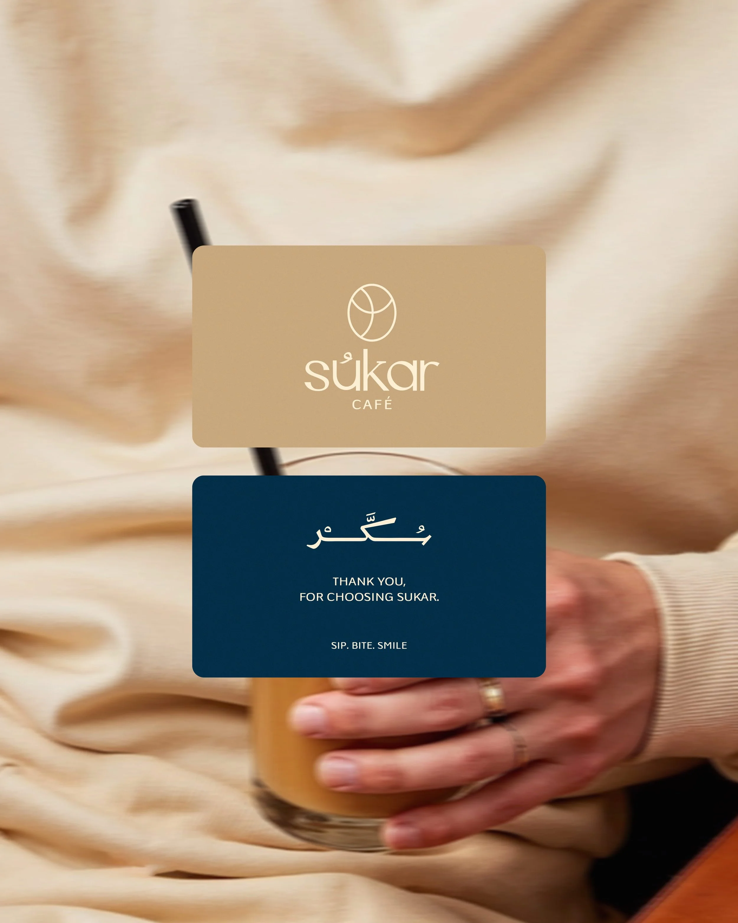 Close-up of a hand holding a cup of coffee with a branded sleeve from Sukar Café. Two cards are partially visible, one beige with the Sukar Café logo and name, and another dark blue with the message in Arabic and English reading, 'Thank you for choos