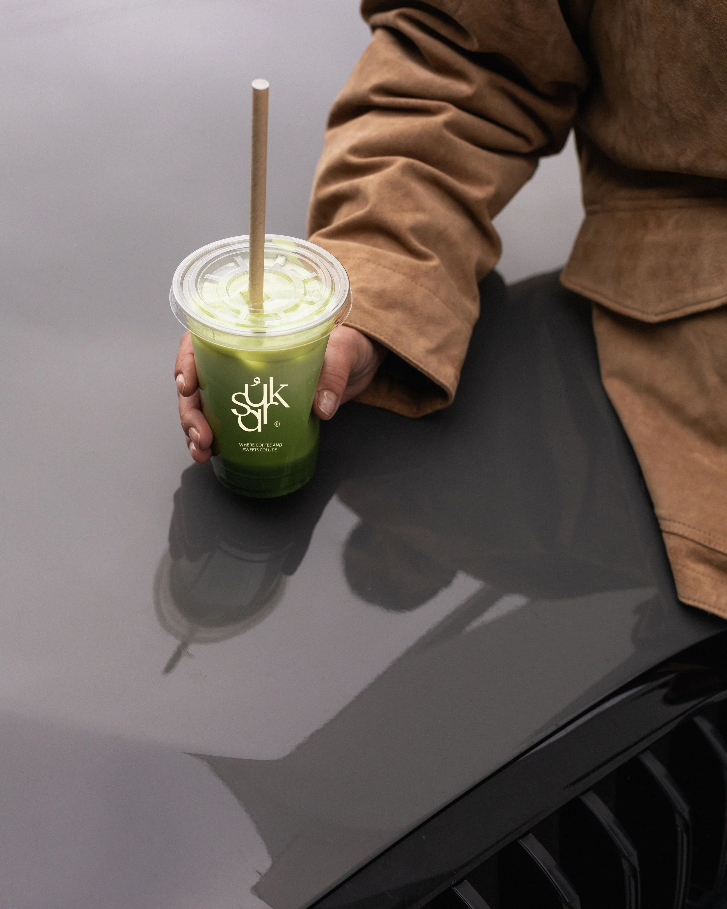 Person holding a green drink with a straw, sitting at a dark gray table, wearing a brown jacket and black pants.
