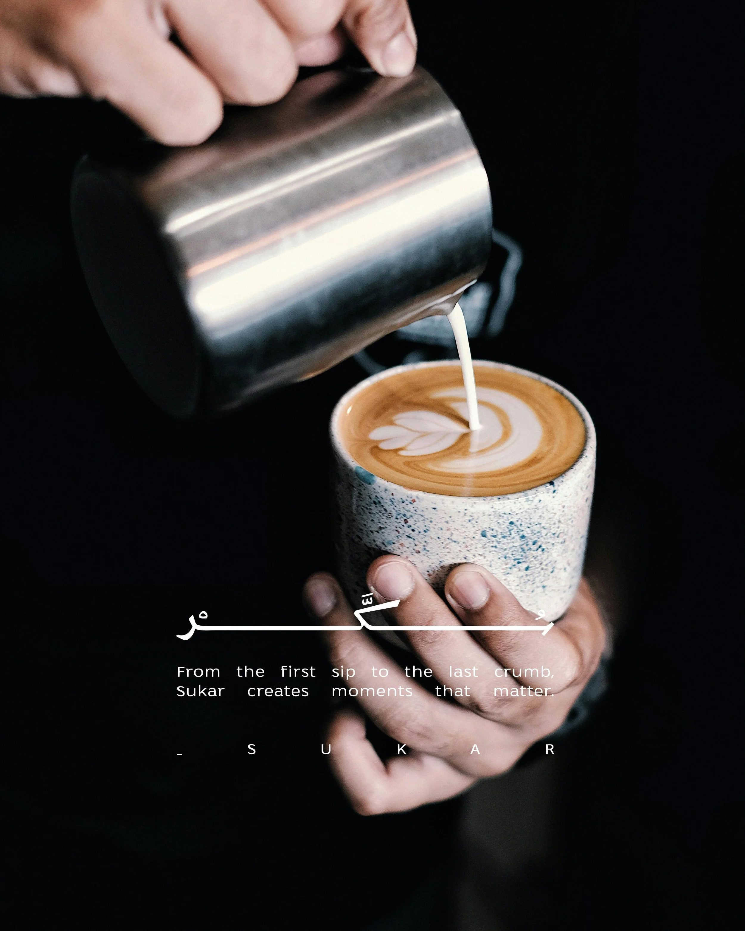A person is pouring steamed milk from a metal pitcher into a cup of coffee to create latte art. The cup is ceramic with a speckled glaze, held by the person's hand, and shows a heart-shaped design in the foam. The background is black, emphasizing the
