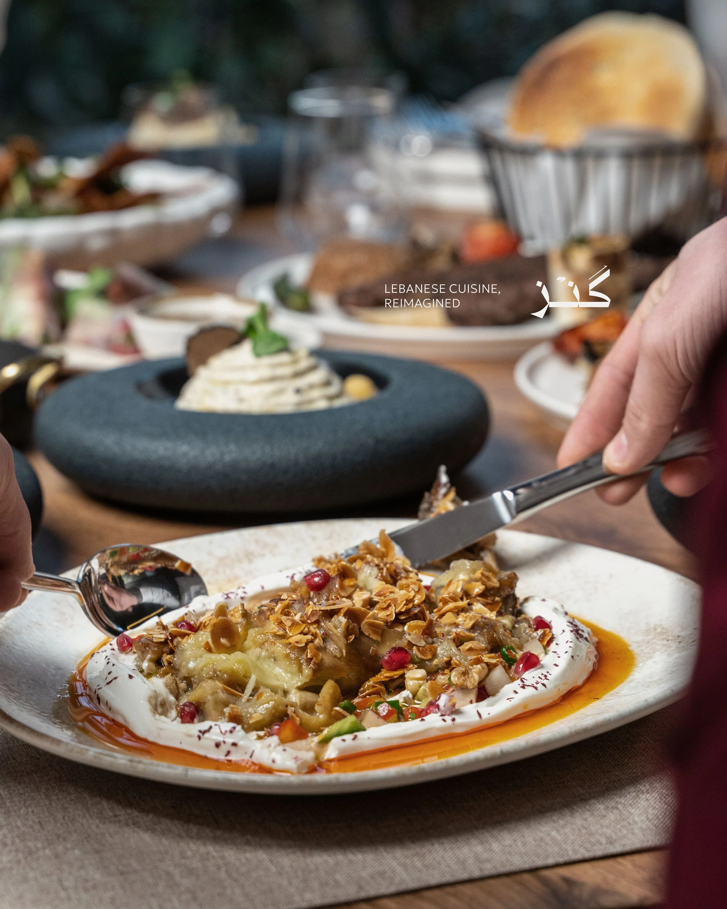 A plate with a Lebanese dessert topped with nuts and pomegranate seeds, surrounded by syrup, on a table with a beige tablecloth, with other food dishes in the background.