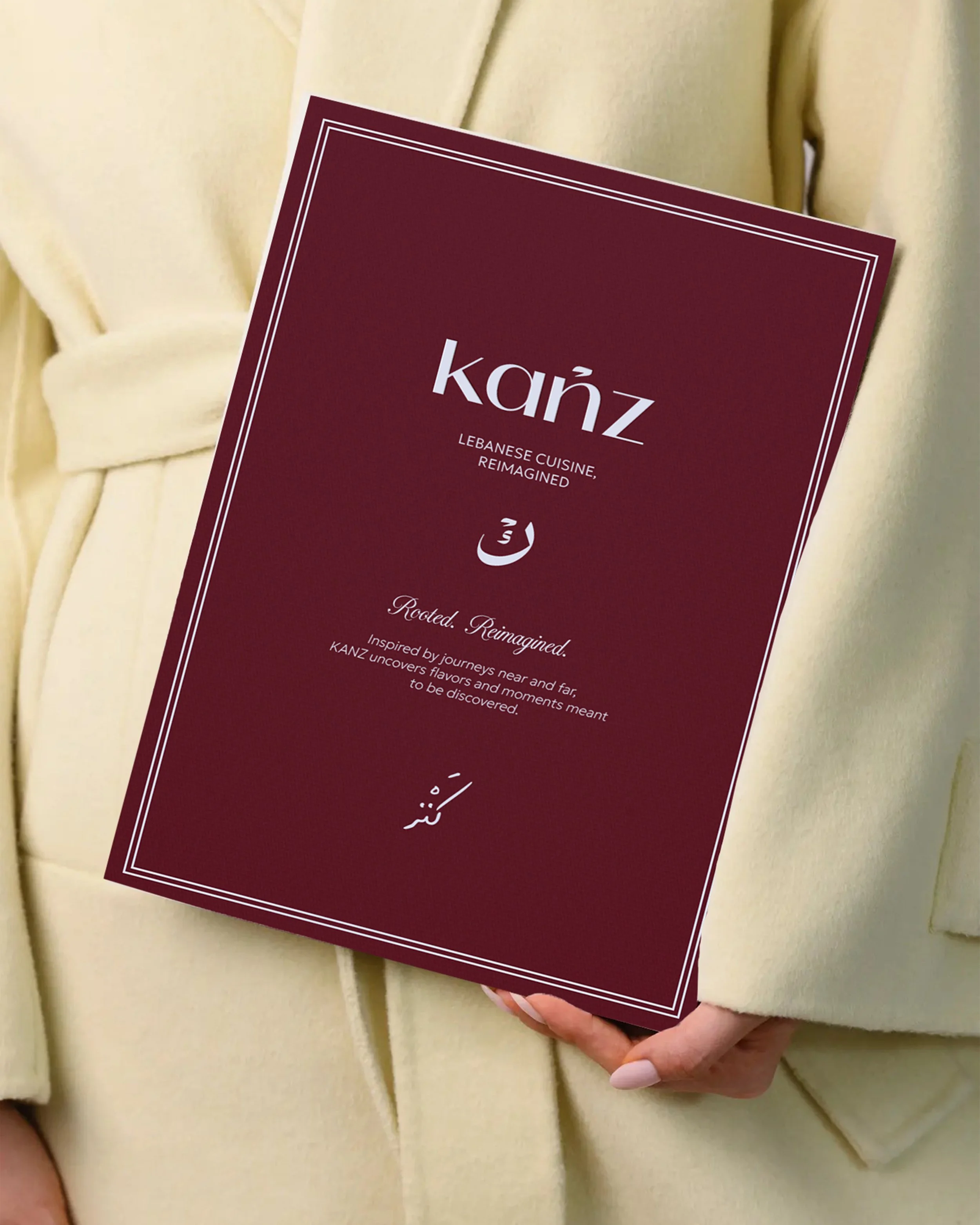 Person holding maroon booklet titled 'kāz' with white text and border, wearing light yellow coat, standing against a cream-colored wall.