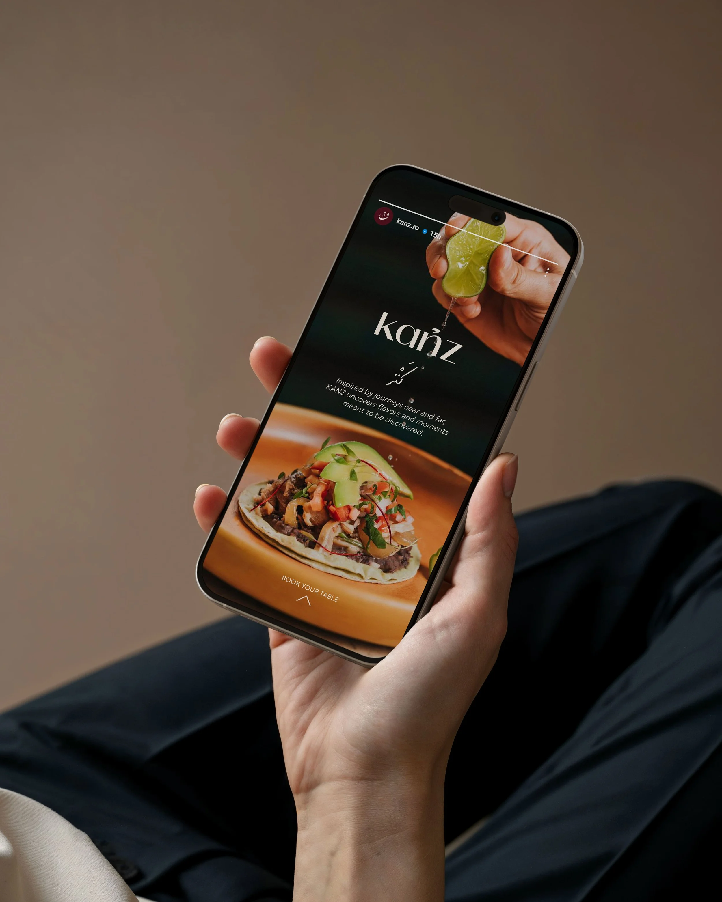 A person holding a smartphone displaying a website for Kanz, a restaurant or food service, with images of a lime and a taco. The phone is held in the person's left hand while seated, with their legs partially visible.