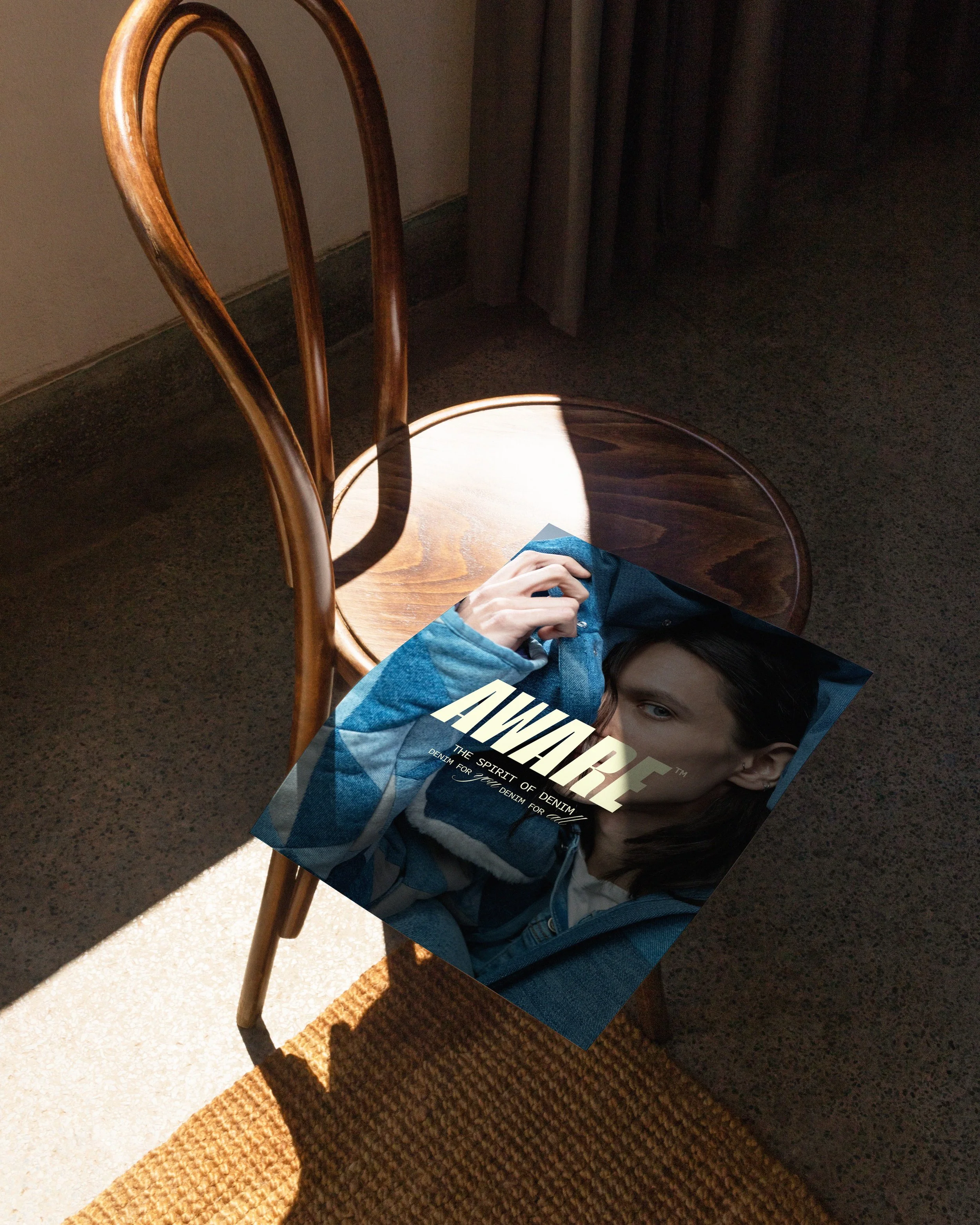 A wooden chair with a rounded backrest, partially illuminated by sunlight, is positioned near a wall with a curtain. A person wearing a blue denim jacket is lying under the chair, holding a magazine titled 'AWAKE' featuring a woman's face on the cove