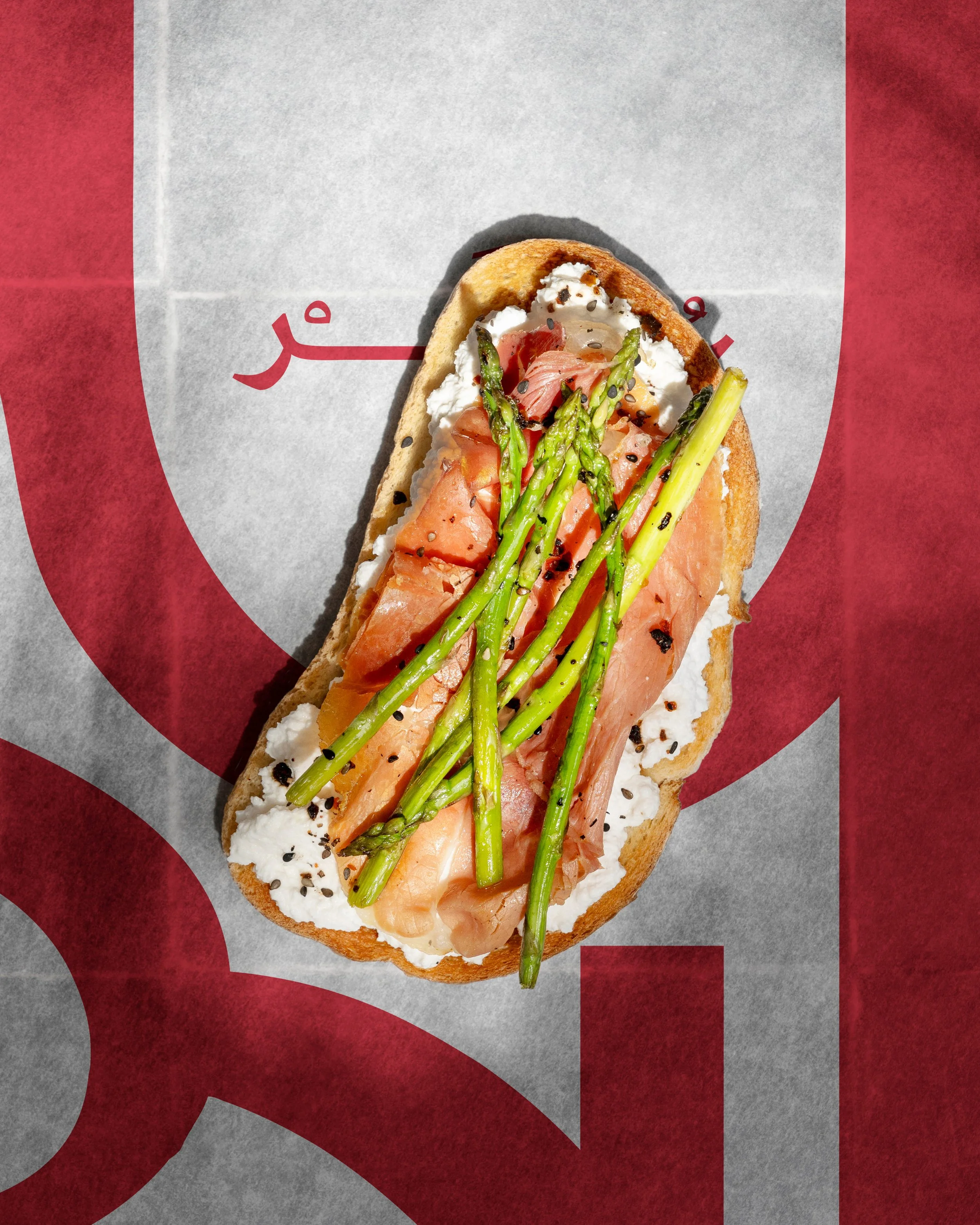 Open-faced sandwich on a baguette slice topped with cream cheese, smoked salmon, grilled asparagus, and black sesame seeds, placed on red and white paper with indistinct red Arabic script.