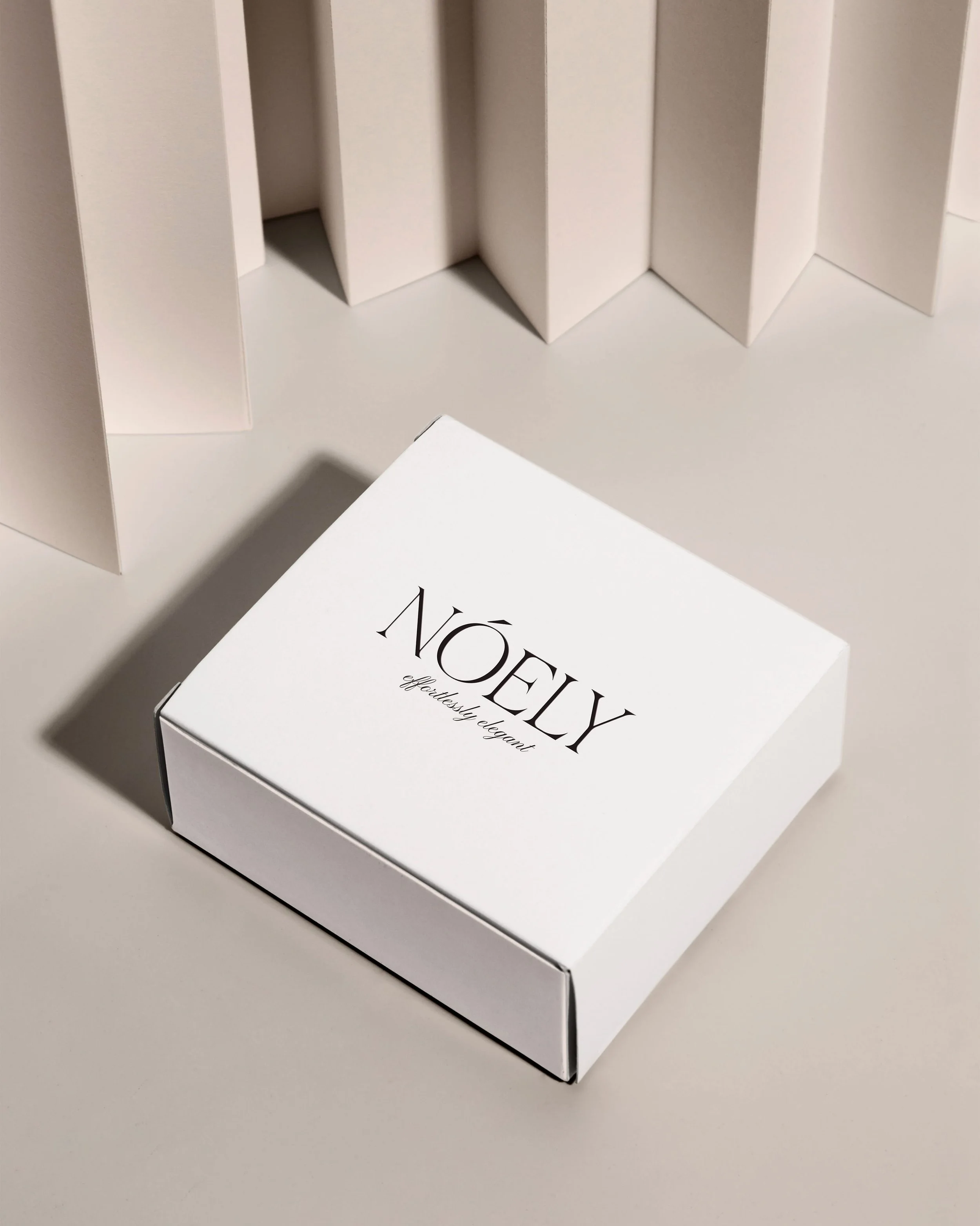 A white box labeled 'NOELY' with the subtitle 'effortlessly elegant' on a white surface, with white paper sheets behind it.