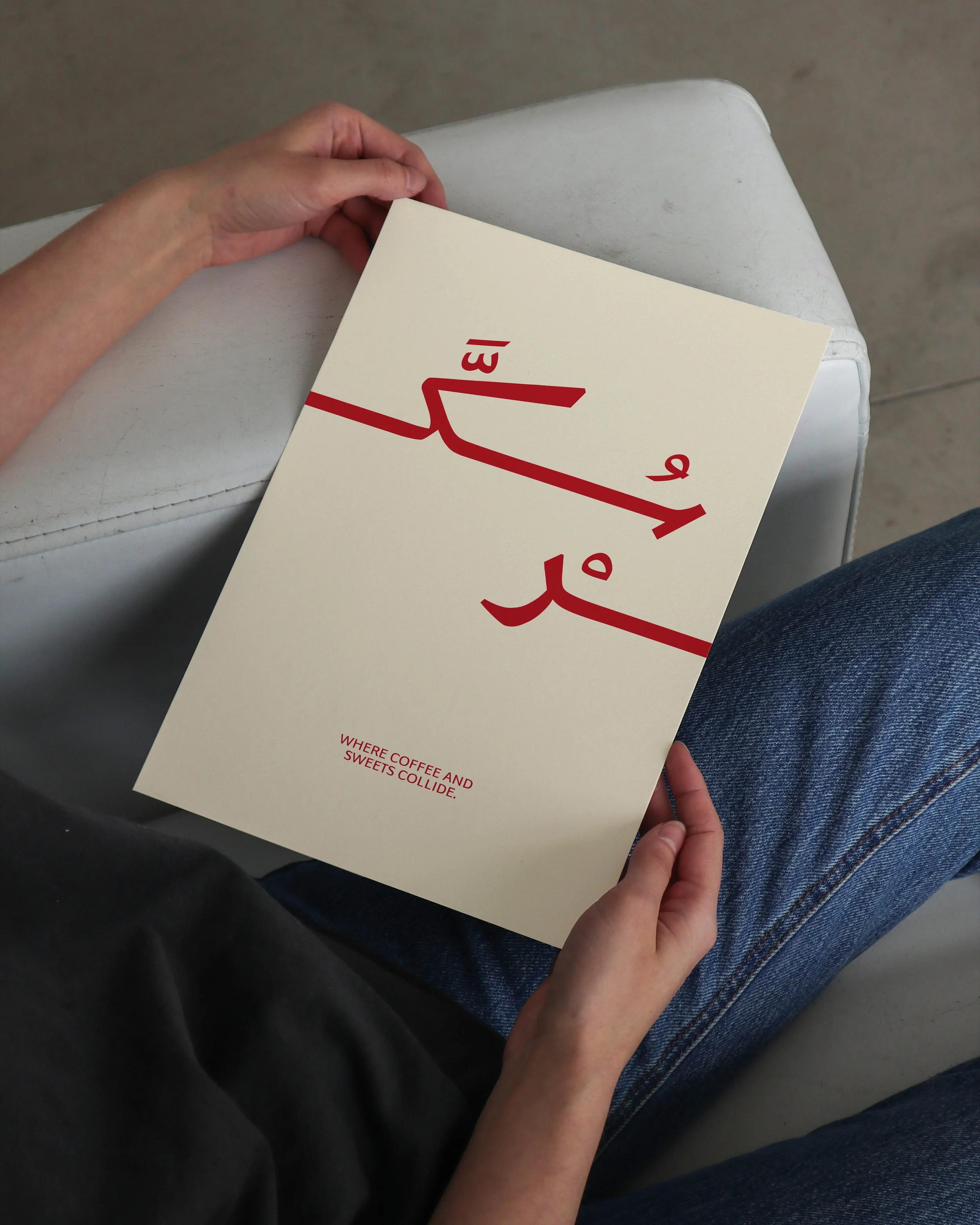 A person holding a white paper with red text and a red graphic, seated on a white chair, wearing blue jeans, with dark floor visible.
