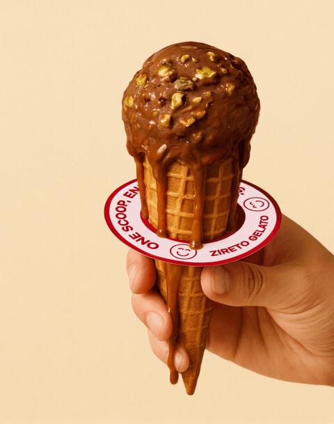 Ice cream cone with chocolate ice cream topped with nuts, held by a hand against a beige background.