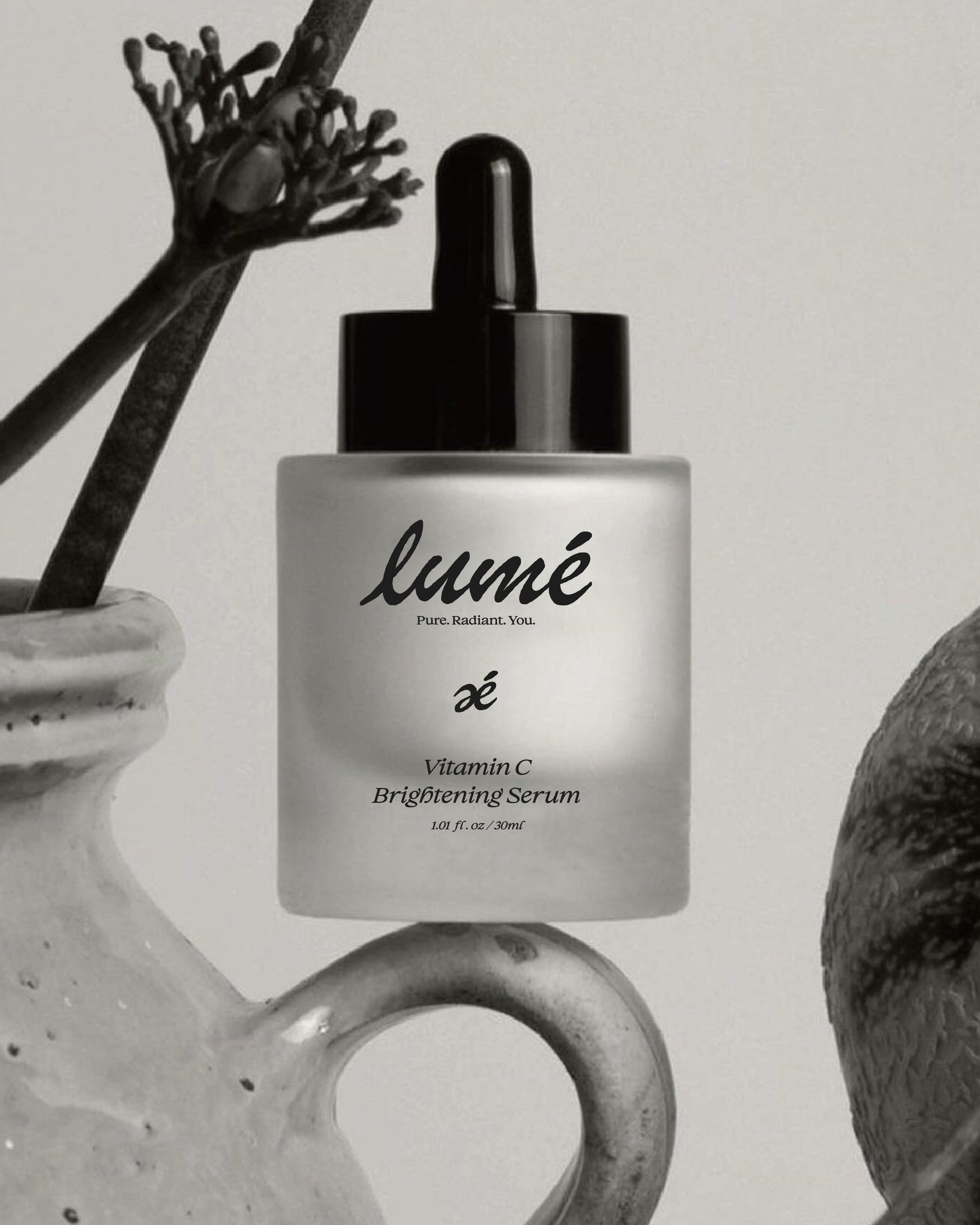 Close-up of a white bottle labeled 'lumé' with a black cap, alongside a black dropper bottle labeled 'é', positioned among smooth, rounded stones and a white marble container.