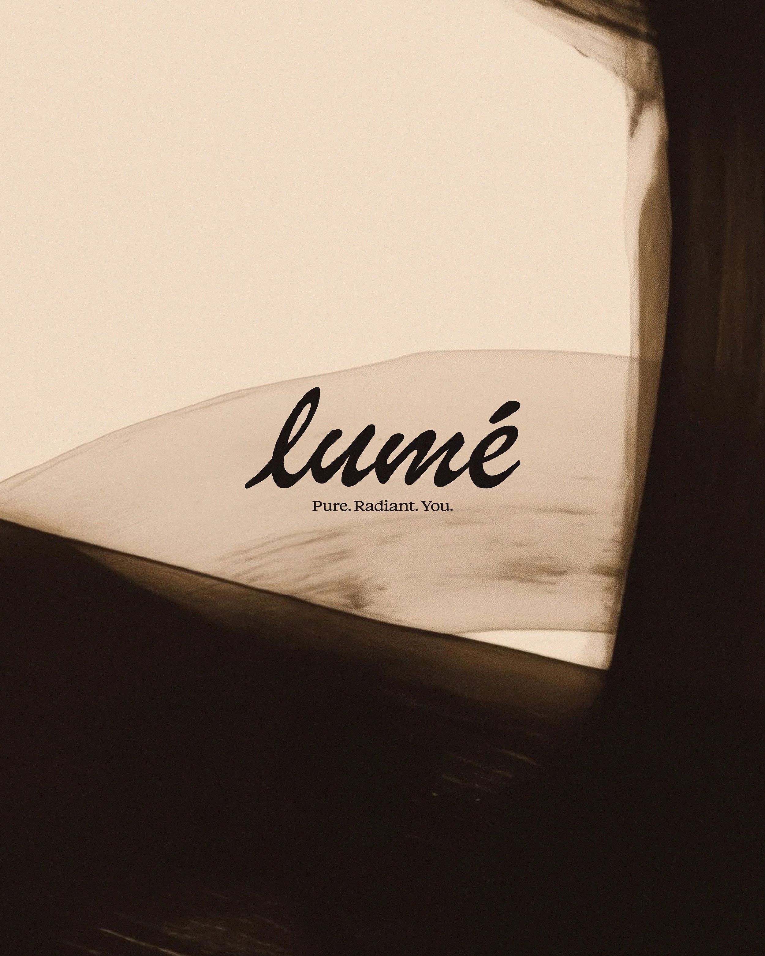 Close-up of a white box with black cursive text stating 'lumié' and a tagline underneath reading 'Pure. Radiant. You.' The box is placed on a dark surface with soft lighting.