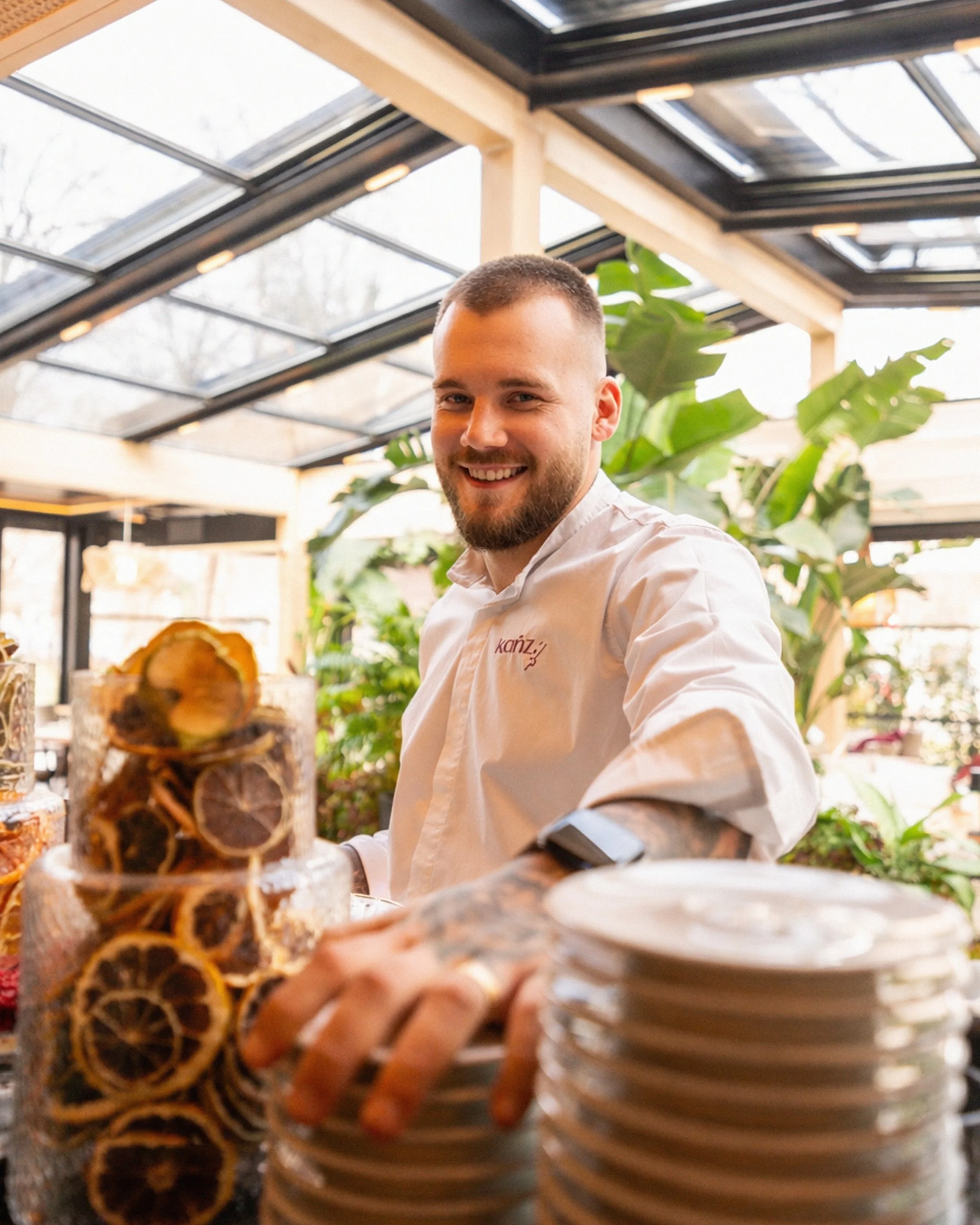 A smiling man with a beard and short hair wearing a white shirt, standing in a bright indoor space with large windows and green plants, with a tower of logs and plates of food in the foreground.