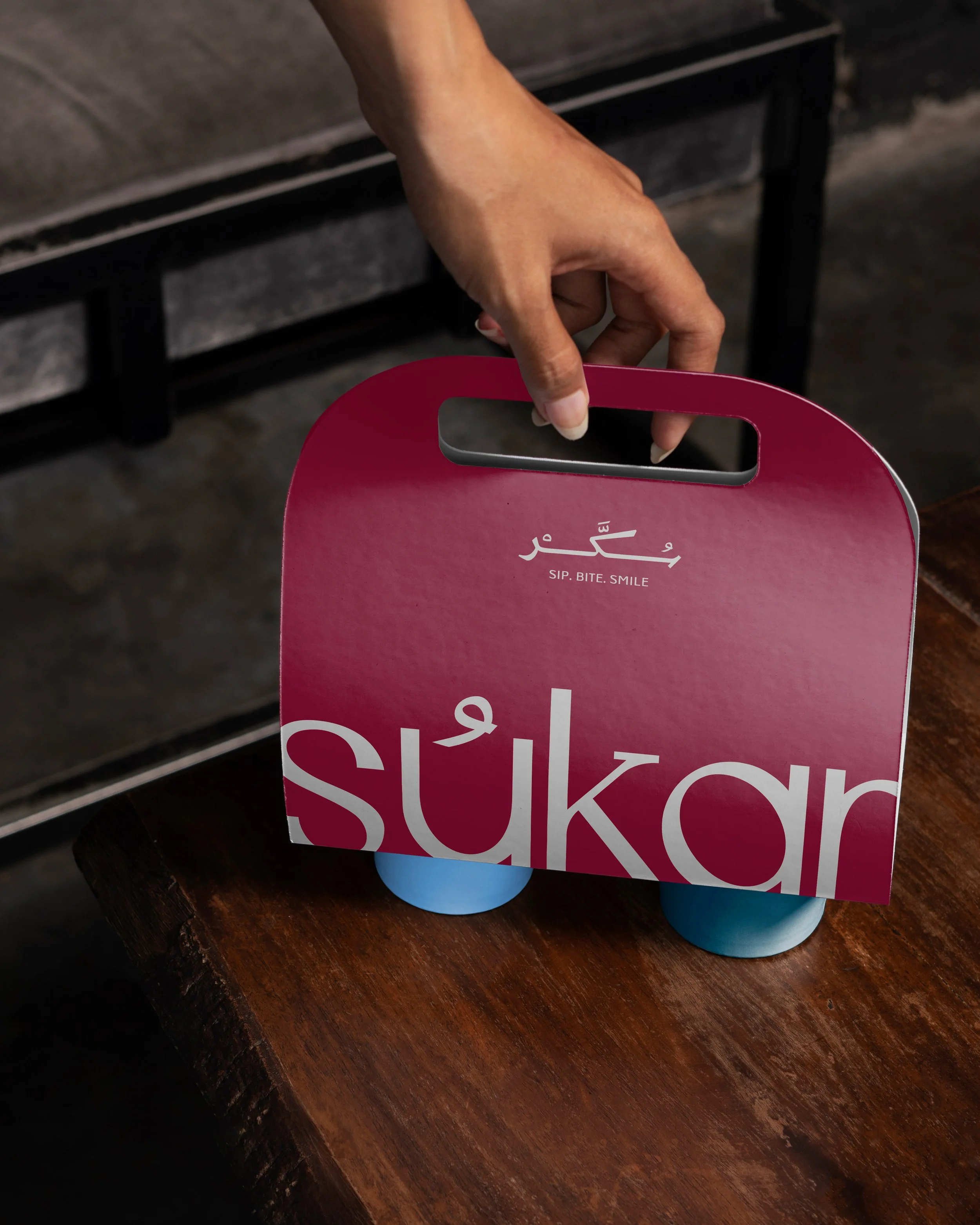 A pink box with Arabic and English text partially visible, held by a person's hand, resting on a dark wooden surface, with a blue object underneath.