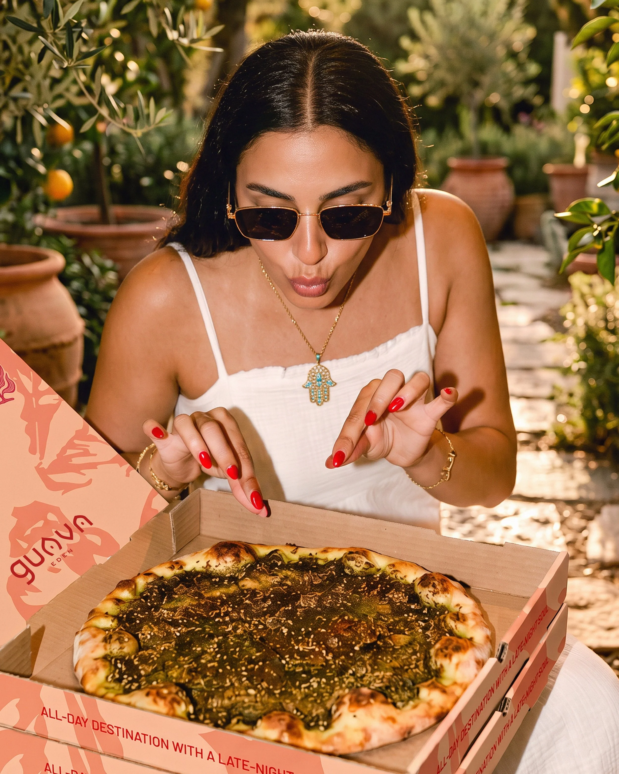 A woman with black hair, wearing sunglasses and a white tank top, is looking at a pizza inside a cardboard box. The background features potted plants and a sunny outdoor setting.