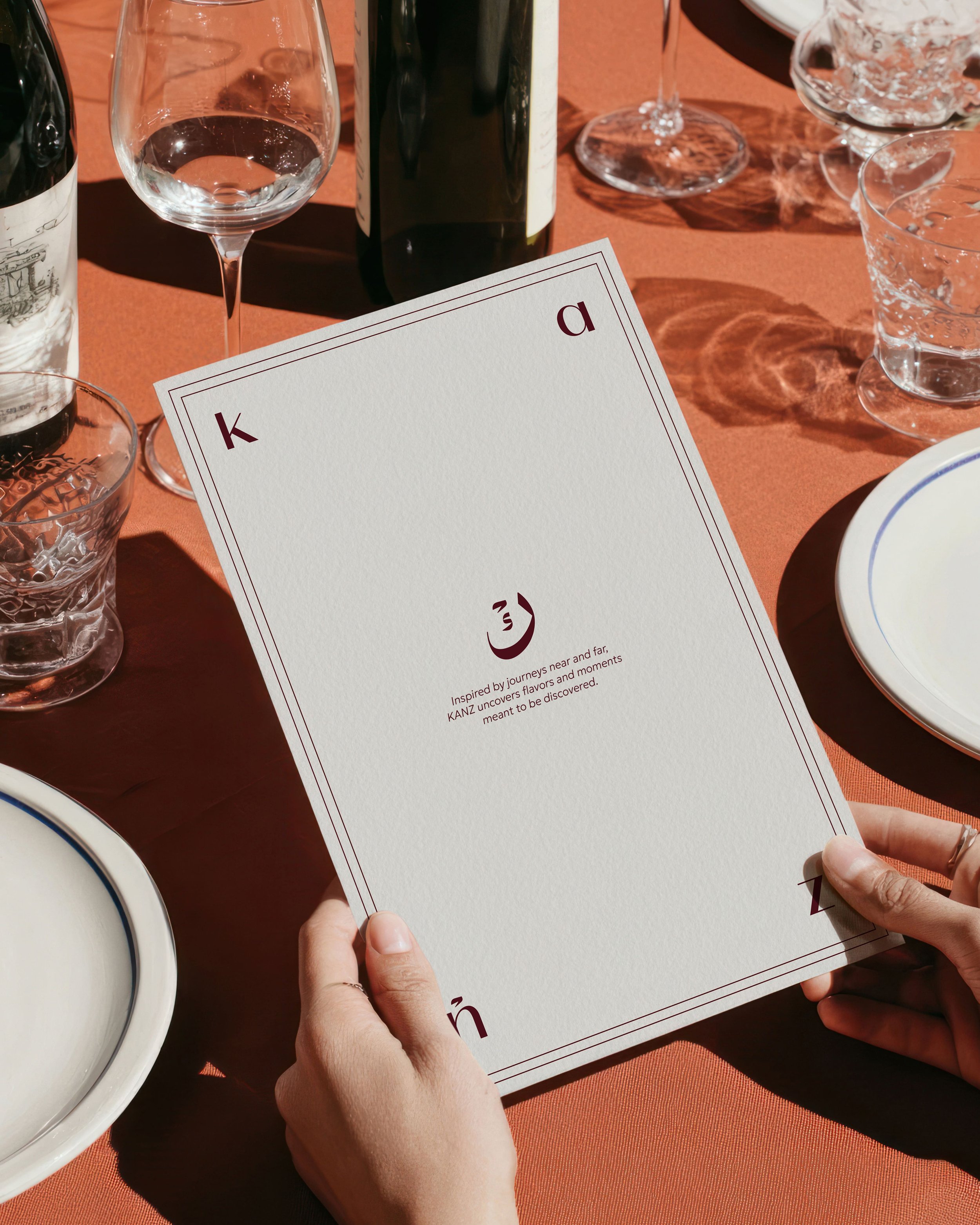 Person holding a white card with the words 'King,' 'Ace,' and 'Queen' printed on it, with other people sitting at a table with wine glasses, a bottle of wine, and plates.