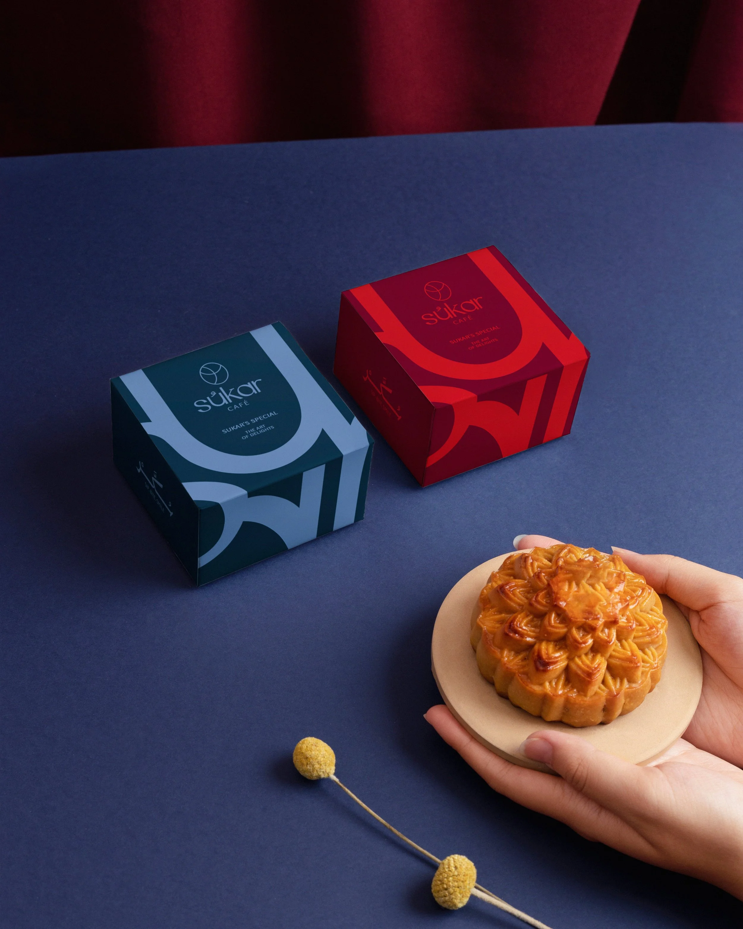 A hand holding a round pastry topped with caramel and noodles on a small beige plate. In the background, there are two boxes labeled Sukar Cafe, one blue and one red, on a dark blue surface. There is also a small yellow pom-pom on a toothpick in the 