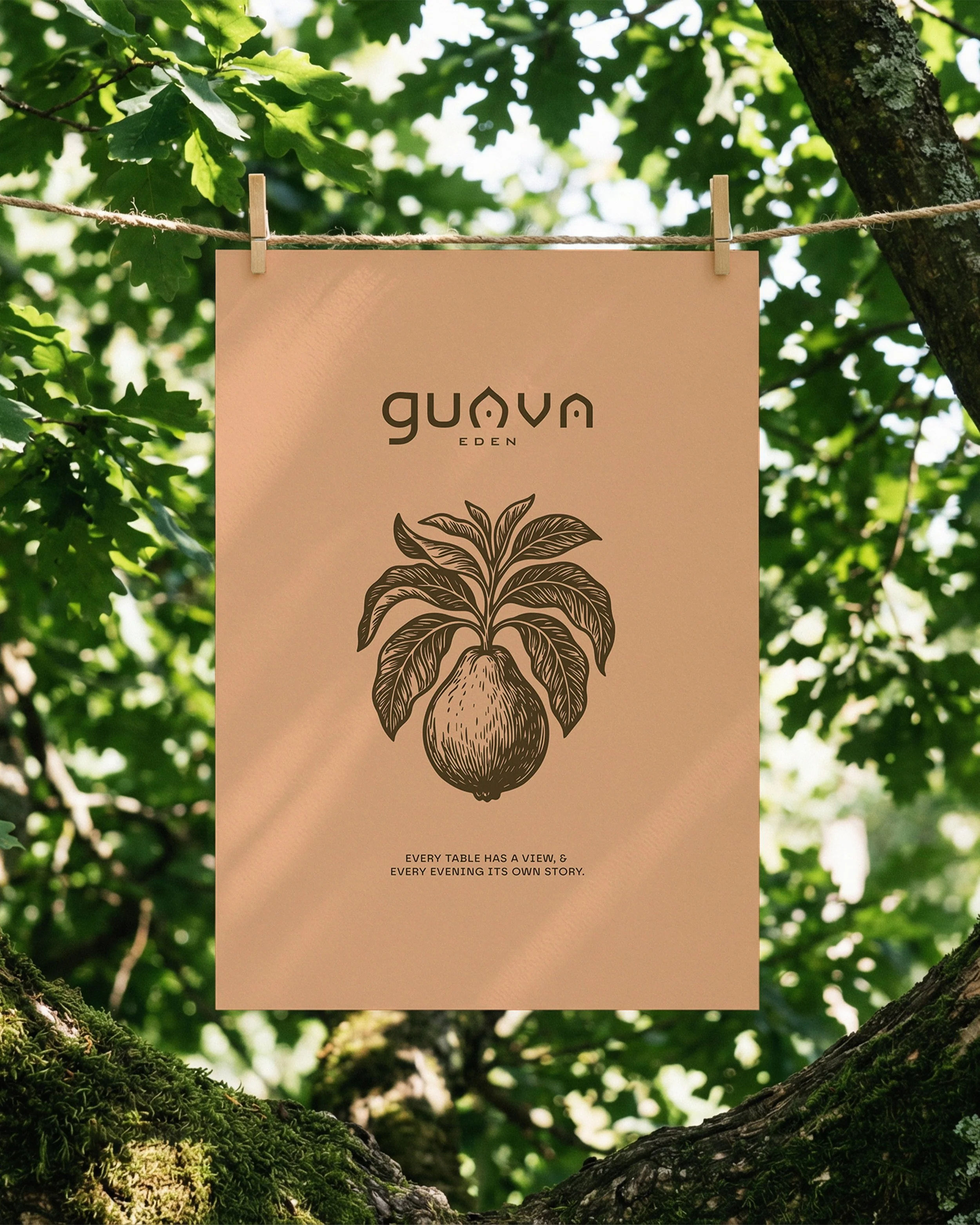 A beige-colored poster hanging on a clothesline between two trees with green leaves. The poster has the words "guava EDEN" and a line drawing of a guava fruit with leaves. Beneath the drawing, a quote reads, "Every table has a view, & every evening i
