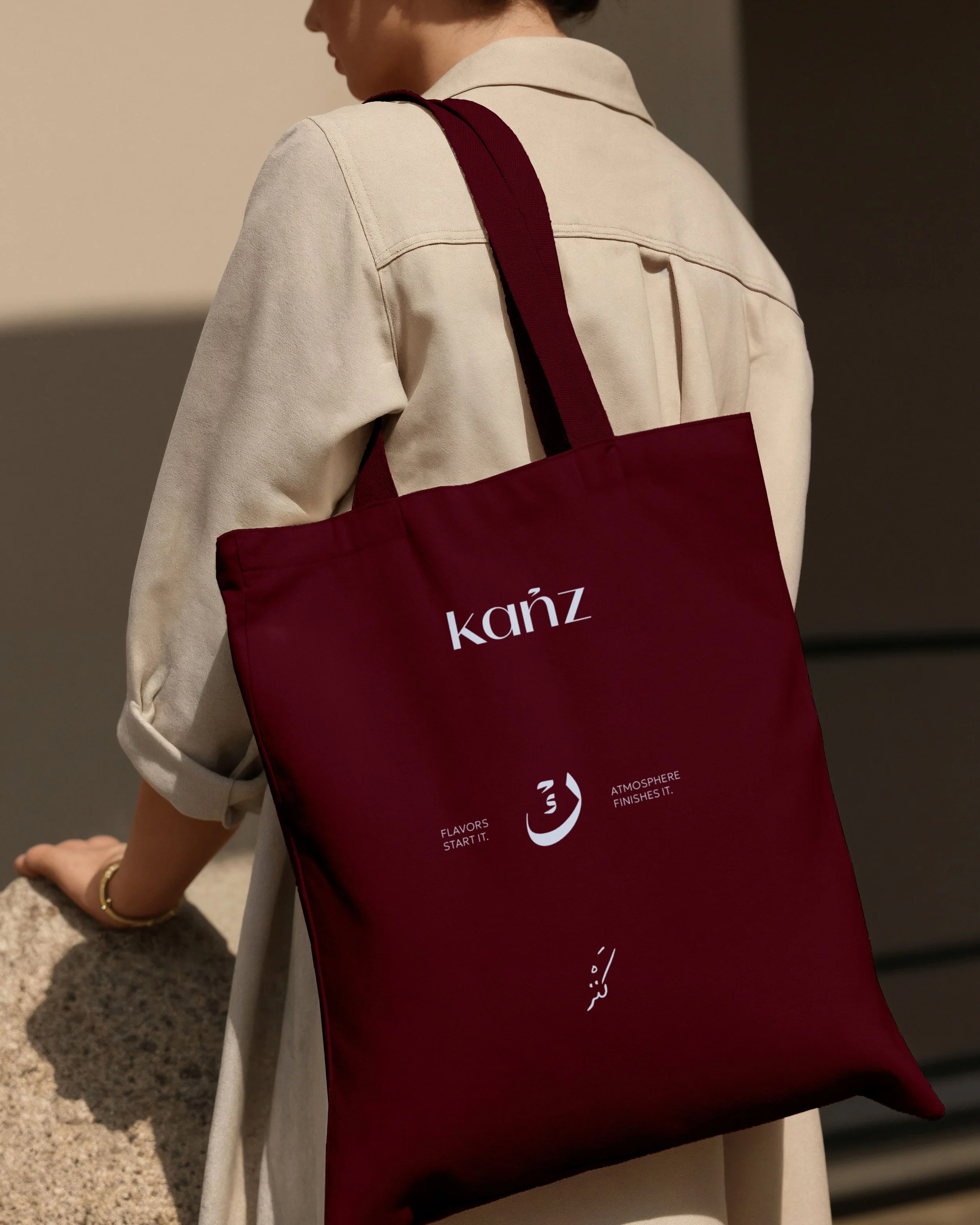 A person wearing a beige blazer with rolled-up sleeves, carrying a maroon tote bag with white text and symbols, standing indoors next to a textured surface.