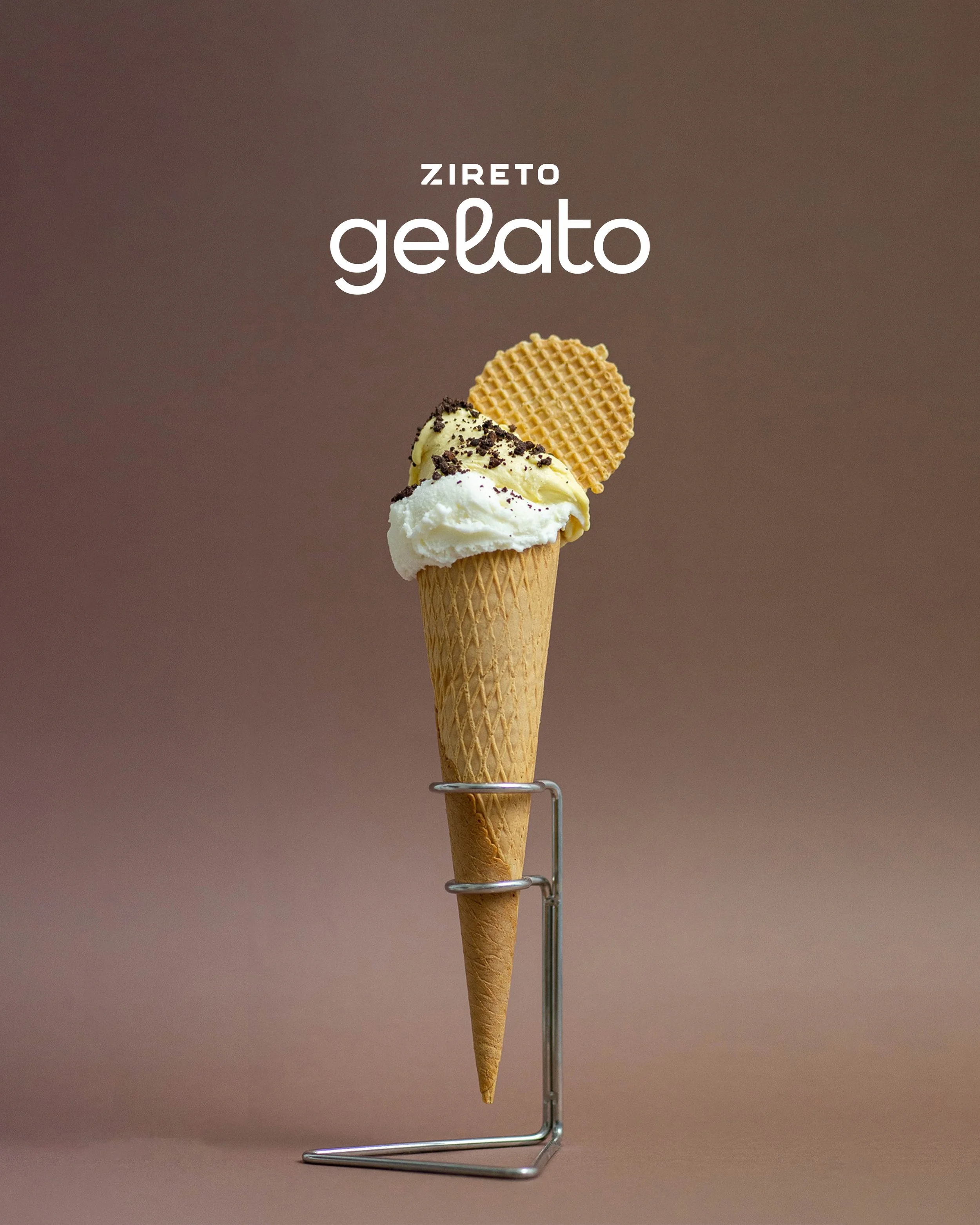 Ice cream cone with vanilla and chocolate topping, and a wafer cookie, displayed on a metal stand with a blurred brown background.