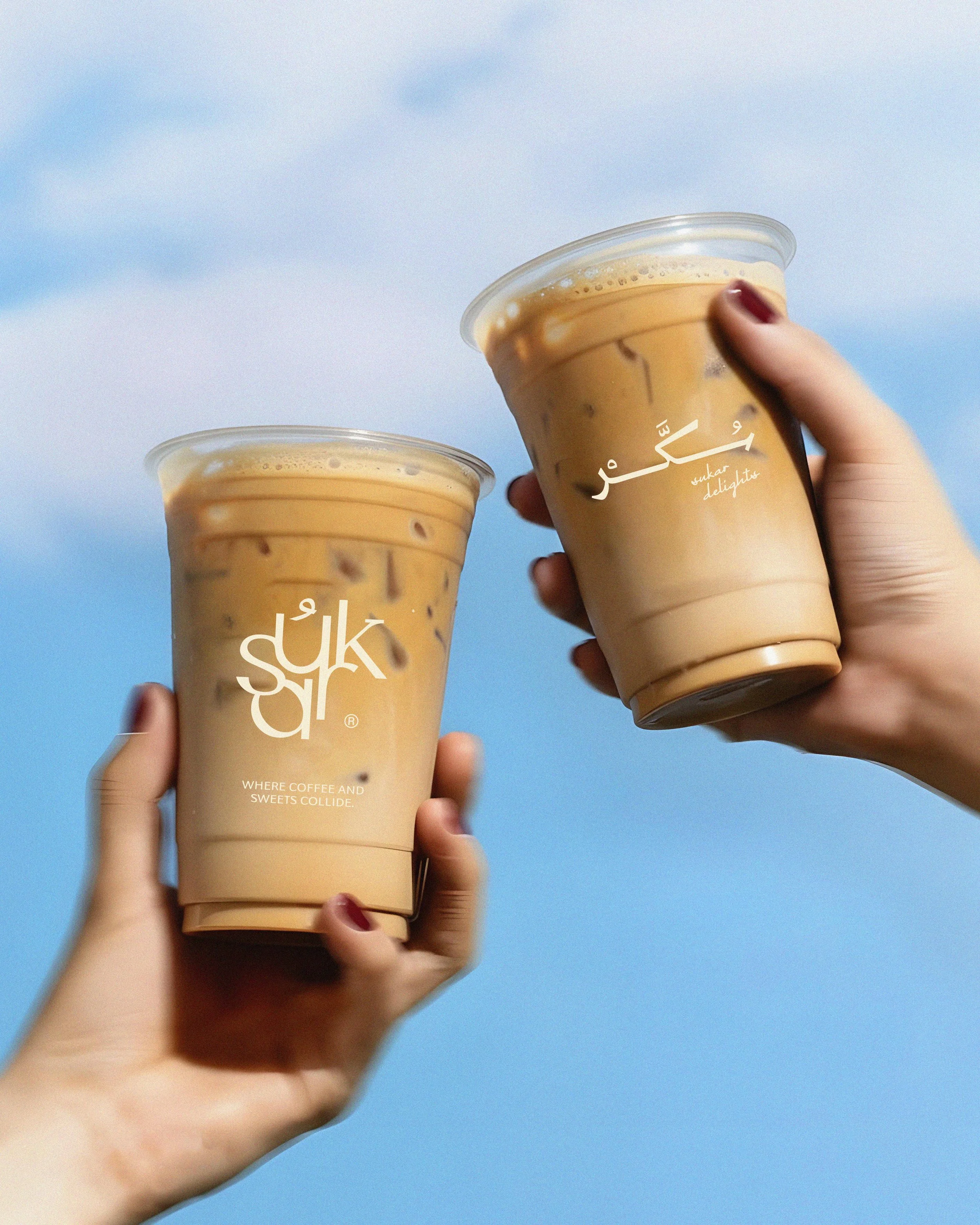 Close-up of a person holding two iced coffee drinks in clear plastic cups with beige lids against a blue sky background.