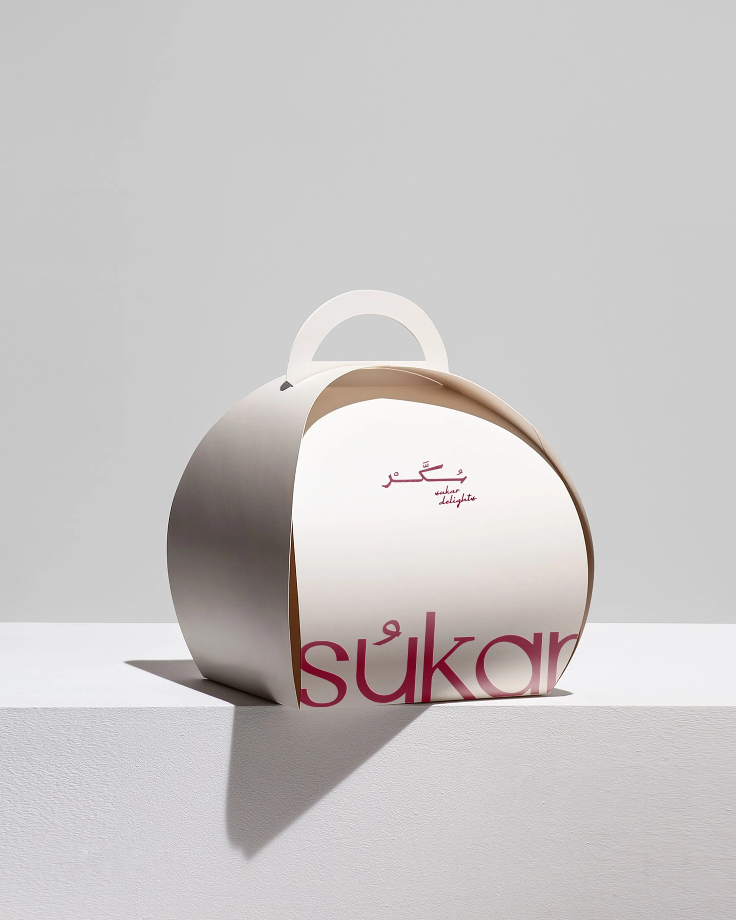 A white paper bag with the brand name Sukar printed in pink, sitting on a white surface with a plain grey background.