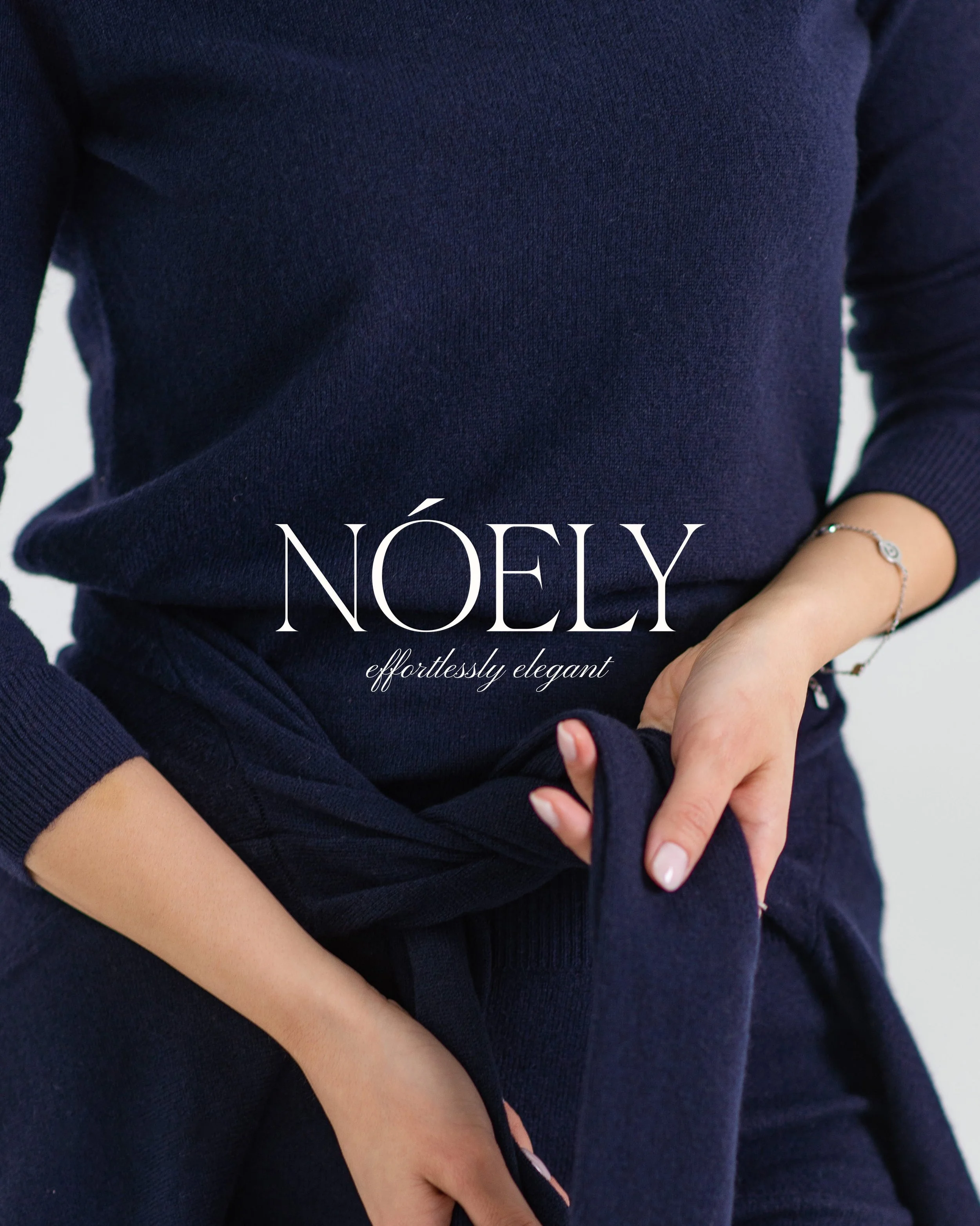 Close-up of a person wearing a dark blue, long-sleeve sweater, with hands crossed over their lap, one hand visible with a delicate bracelet, and the word 'NÖLY' partially visible at the bottom.
