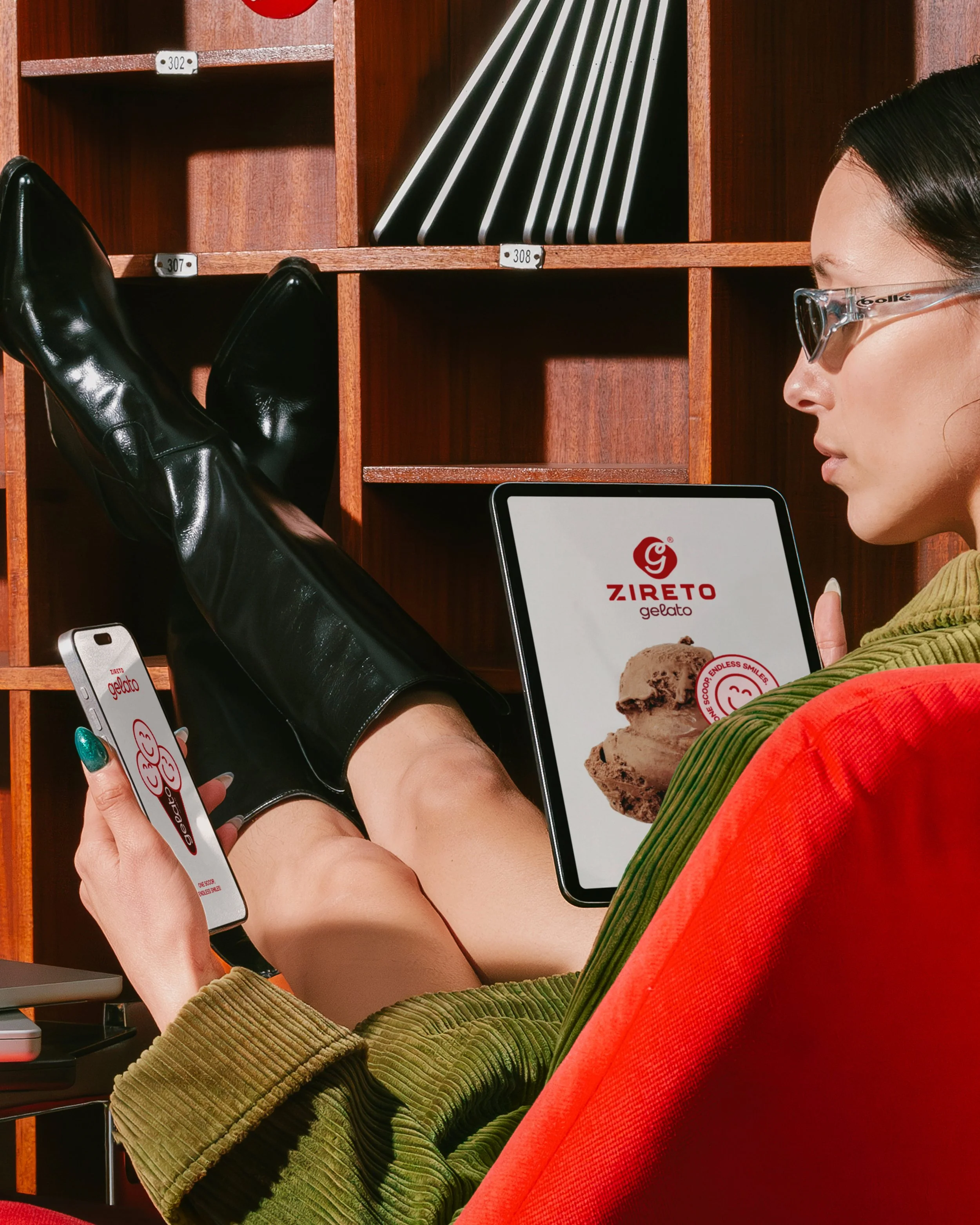 Person wearing glasses and green corduroy jacket, holding a smartphone with 'Zireto Gelato' advertisement, leaning against a wooden cubby shelf.