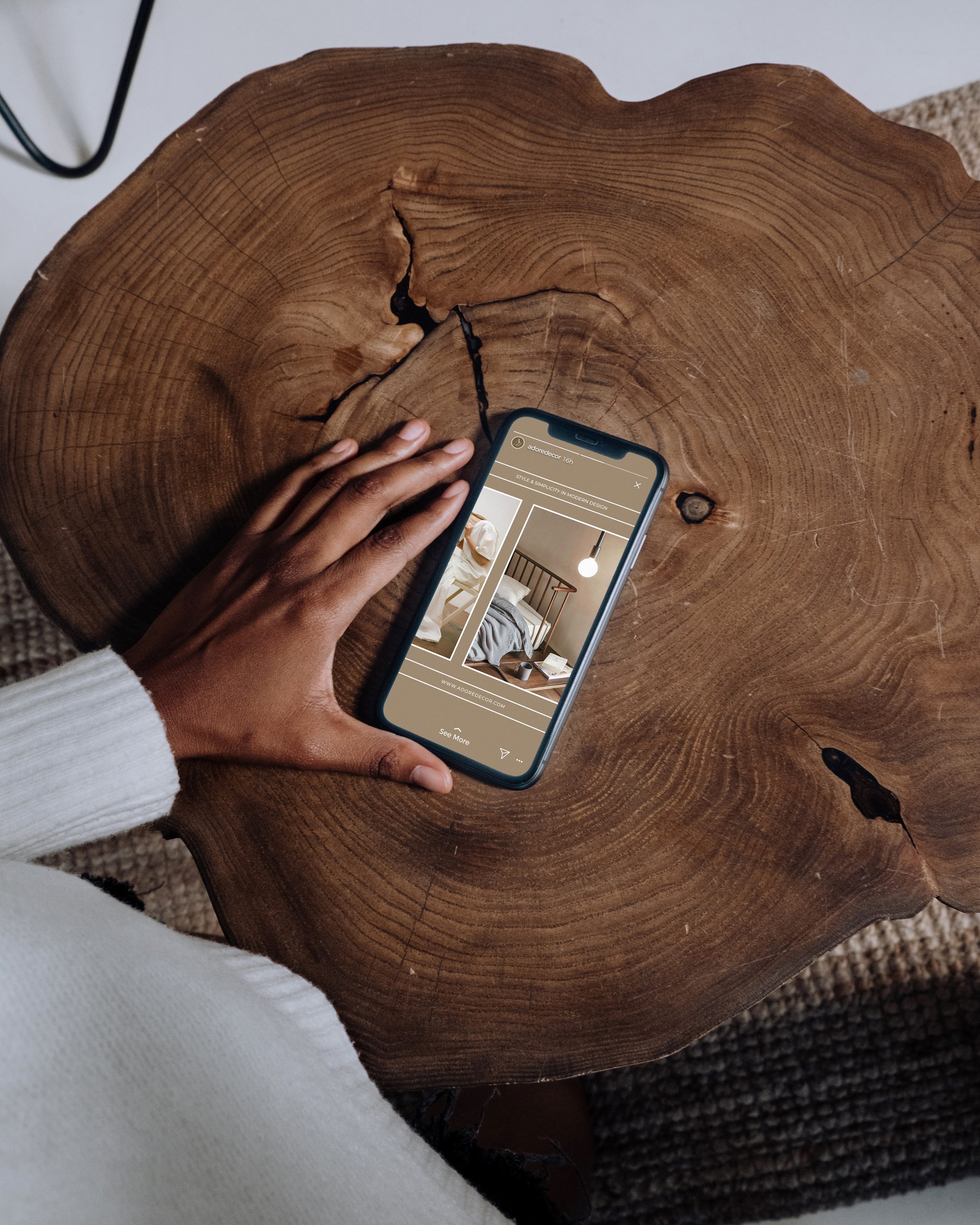 Close-up of a large, cross-sectioned tree trunk with visible growth rings and a natural crack, resting on a textured gray surface. Part of a person's hand and white sleeve are visible holding a smartphone.