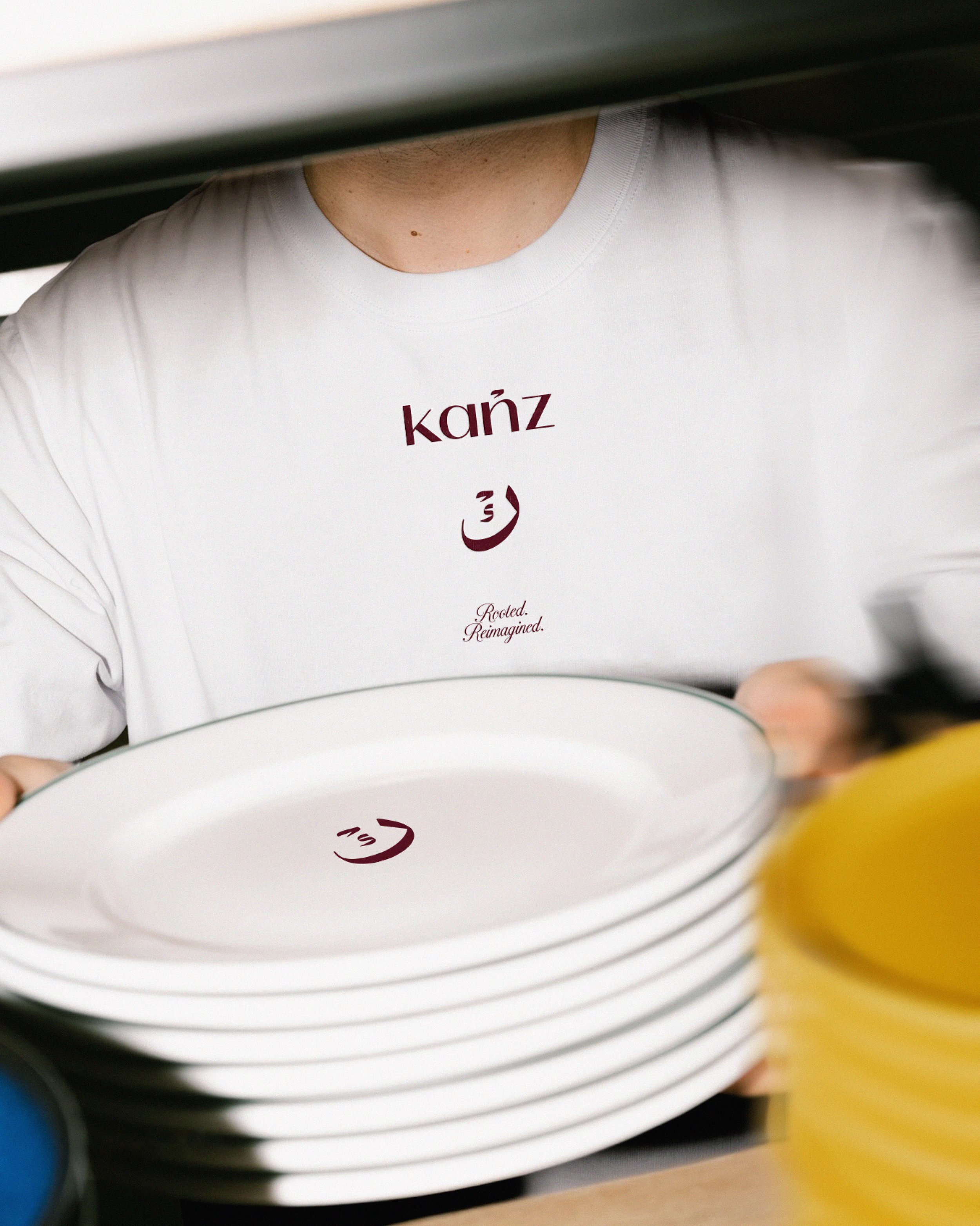 Close-up of a white t-shirt with the word 'kanz' and an Arabic word written in maroon, viewed through a series of stacked plates.